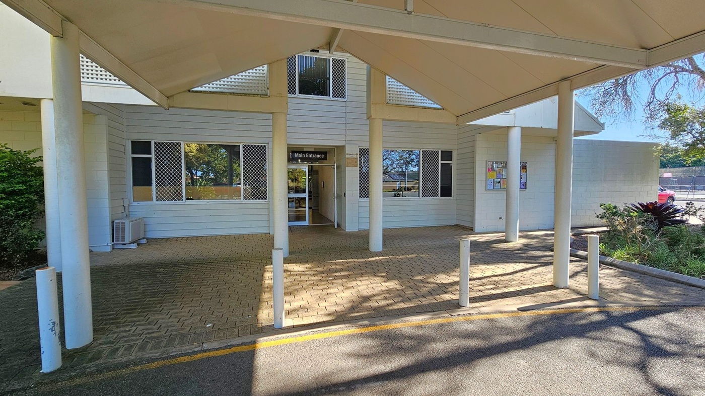 A building entrance with a sign reading "Main Entrance" above a glass door. The structure has light-coloured cladding and several windows with security grilles. A covered walkway supported by white pillars leads to the entrance. The ground is paved with bricks, and there are white bollards in front. On the right, a notice board is mounted on the wall, and a small garden with shrubs is visible. In the background, a car park and trees can be seen.