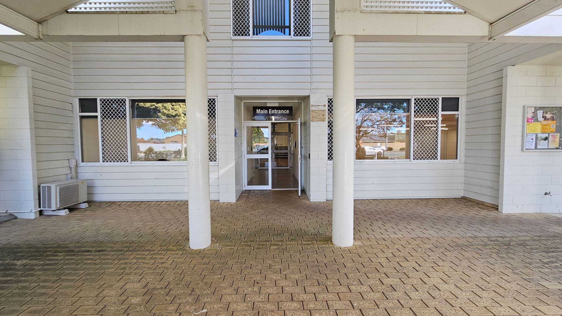 A building entrance with a sign reading "Main Entrance" above glass double doors. The exterior features white horizontal siding and two large windows with security grilles on either side of the doors. An air conditioning unit is visible on the left. The floor is paved with light brown bricks. A notice board with various papers is mounted on the right wall. The ceiling is covered with a light-coloured material, and there are two white columns in front of the entrance.