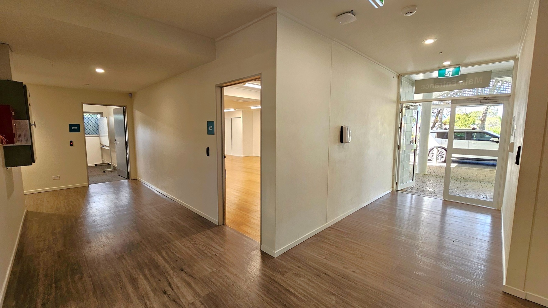 A hallway with wooden flooring and cream-coloured walls. On the left, a door leads to a room with a window and a partially visible sink. In the centre, another door opens to a room with light wooden flooring. On the right, a glass entrance door shows a white car parked outside. The ceiling has recessed lighting and a smoke detector. An exit sign is above the entrance door.