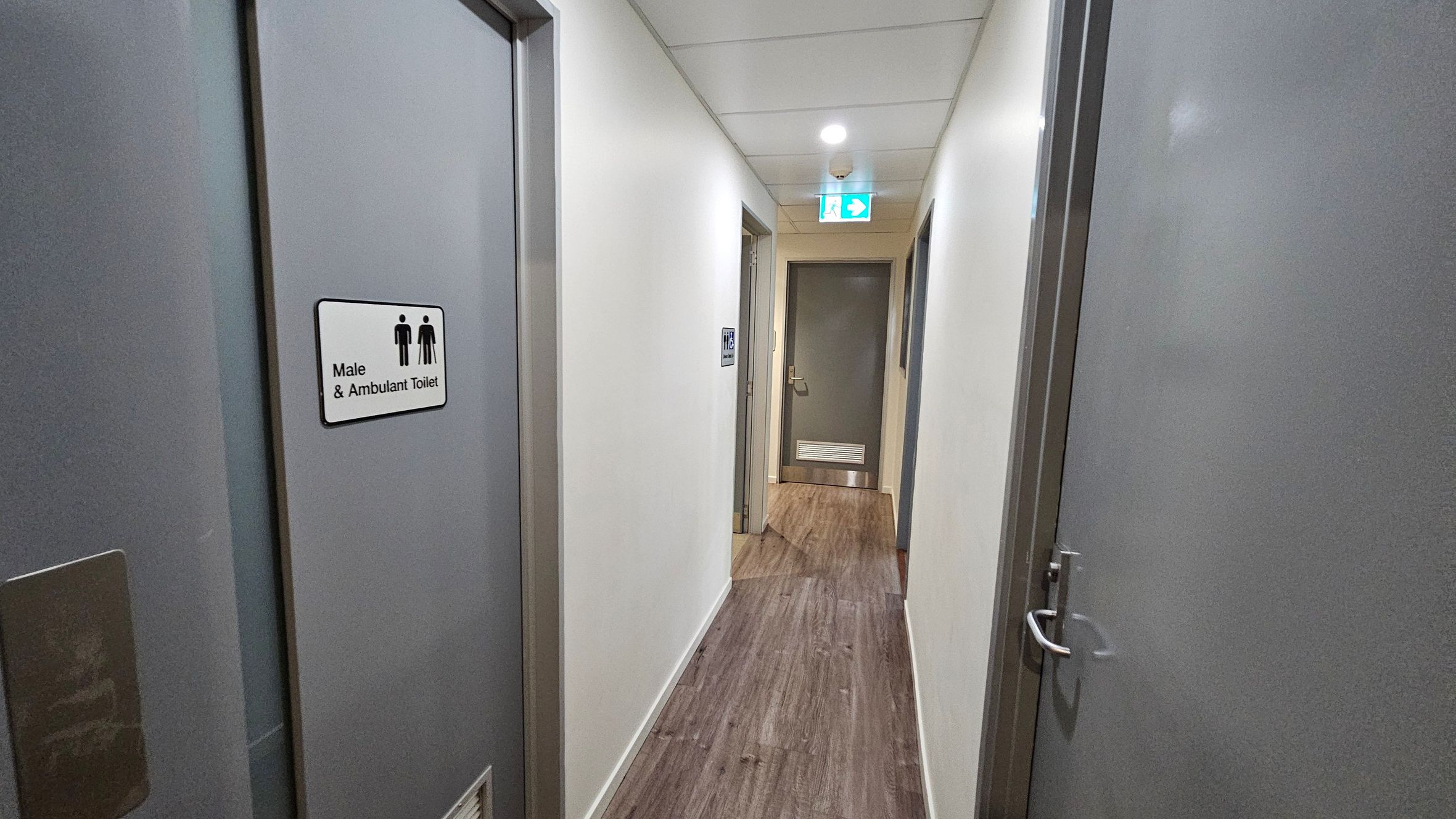 A narrow corridor with white walls and a wooden floor. On the left, a door with a sign reading "Male & Ambulant Toilet" is visible. On the right, another grey door with a handle. At the end of the corridor, a closed grey door with a vent at the bottom. Above, a ceiling with a light fixture and an illuminated exit sign.