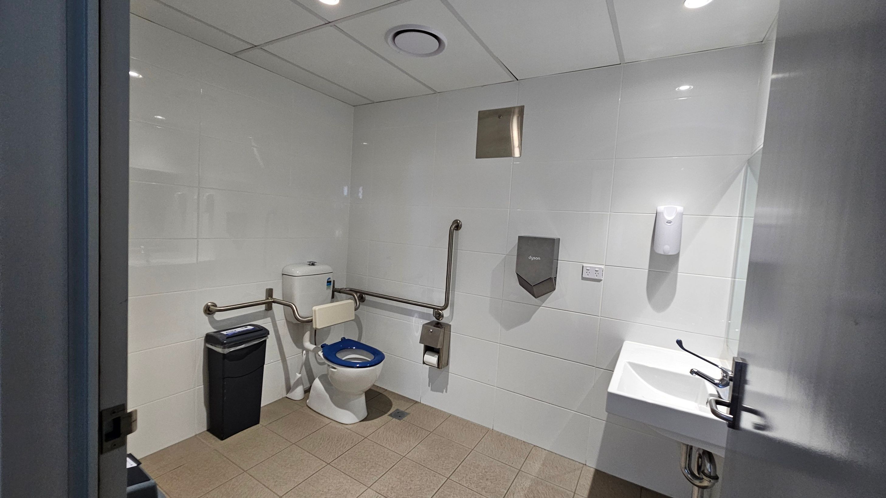 A public restroom with white tiled walls and a beige tiled floor. On the left, there is a toilet with a blue seat, equipped with support rails. A black waste bin is positioned nearby. In the centre, a toilet paper holder and a hand dryer are mounted on the wall. On the right, a white sink with a lever tap is visible, along with a soap dispenser above it. The ceiling is fitted with recessed lighting and a ventilation duct.