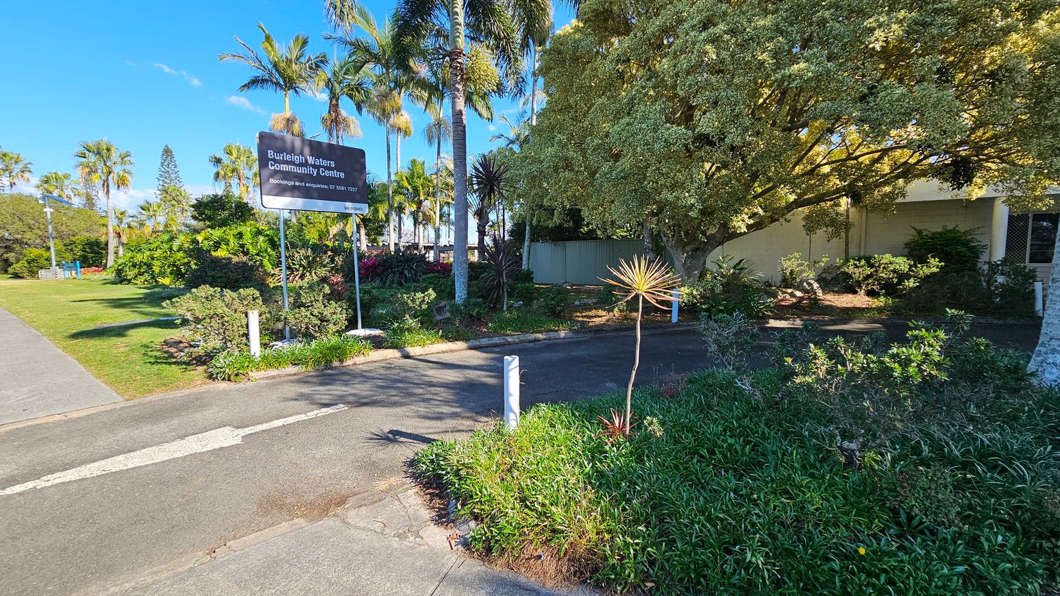 A sign for "Burleigh Waters Community Centre" is on the left, surrounded by lush greenery and palm trees. A paved path runs alongside a grassy area with more trees in the background. On the right, a large tree provides shade over a garden area with various plants. A narrow road or driveway is visible, bordered by low vegetation. The scene is well-lit, suggesting a sunny day.