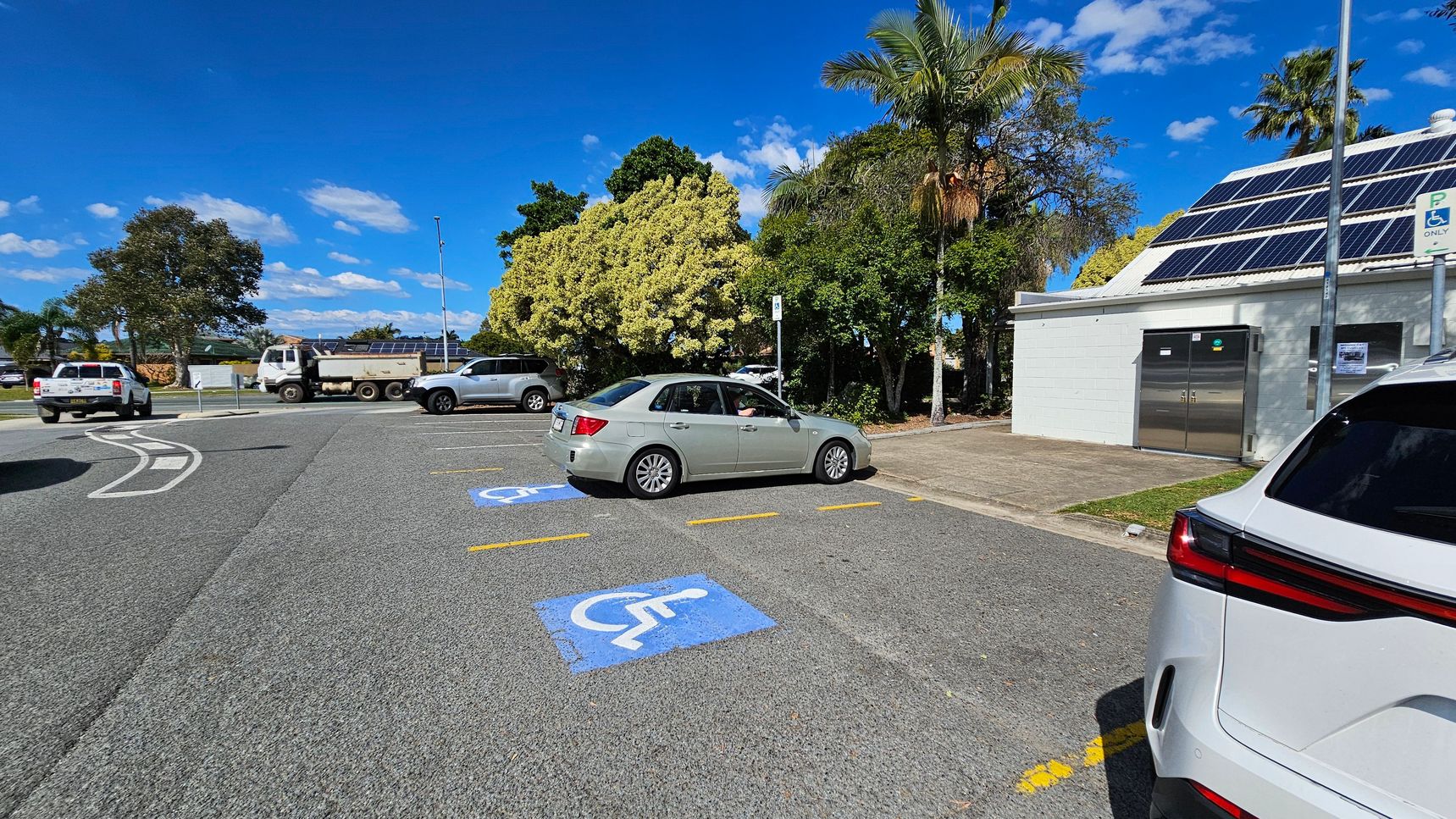 A car park with designated disabled parking spaces marked in blue on the asphalt. On the right, a white building with solar panels on the roof and a metal door. A white SUV is partially visible in the foreground. In the background, there are several trees and a few vehicles, including a truck and a silver car. The area is well-lit, suggesting a sunny day.
