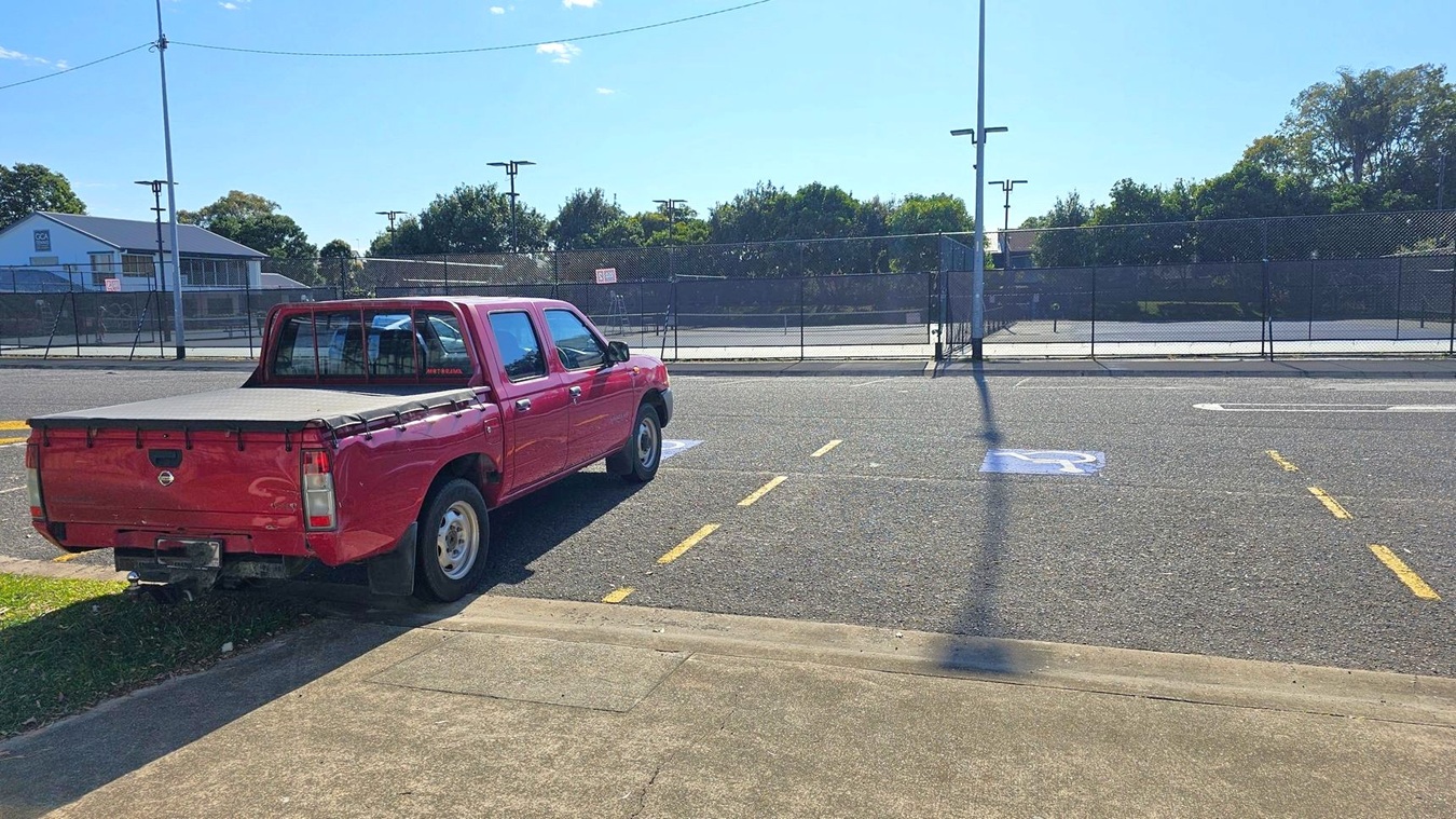 A red pickup truck is parked on the left side of a paved area, partially on the grass. The vehicle is facing away from the camera. In the background, there is a fenced tennis court with trees and a building visible beyond it. The road has a marked disabled parking space and yellow lines. A tall streetlight pole casts a shadow on the ground. The pavement in the foreground is concrete.