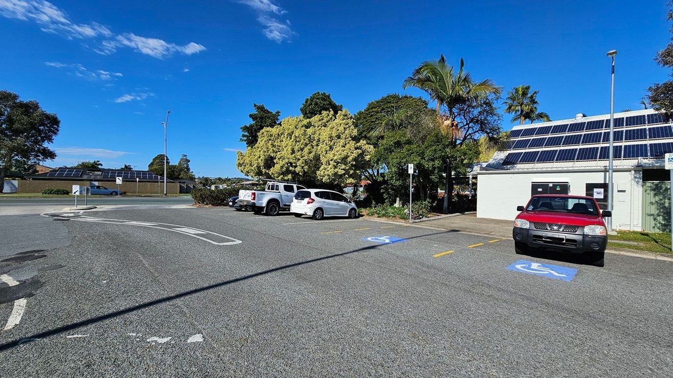 A car park with several vehicles, including a red car in a disabled parking space on the right. A white building with solar panels on the roof is adjacent to the car. Trees with dense foliage are in the background, and a white and a silver vehicle are parked nearby. A road with a car driving past is visible on the left, bordered by a low wall with solar panels. The ground is paved, and a streetlamp is present near the building.