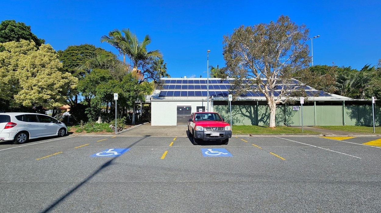 A parking area with a red vehicle parked in a space marked for disabled access. To the left, a white car is parked. The background features a building with solar panels on the roof, surrounded by lush green trees and foliage. The ground is paved with marked parking lines, and there are signs indicating disabled parking. A green fence and a tree are visible to the right of the building.