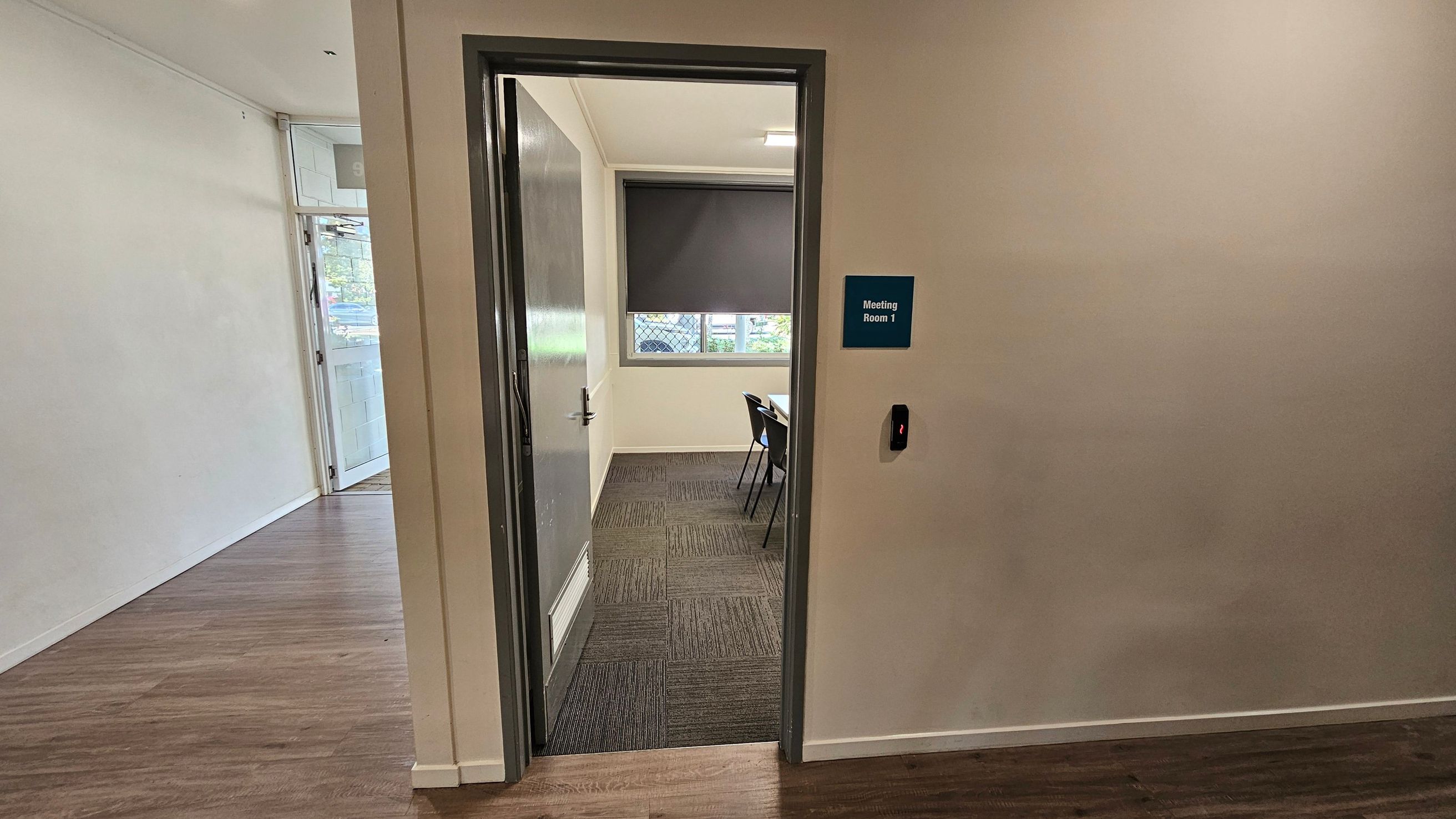 A partially open door leads into a room labelled "Meeting Room 1" on a blue sign. The room has a window with a closed blind and a table with chairs. The floor inside the room is carpeted, while the hallway has a wooden floor. A card reader is mounted on the wall next to the door. The walls are plain, and the ceiling is not visible.