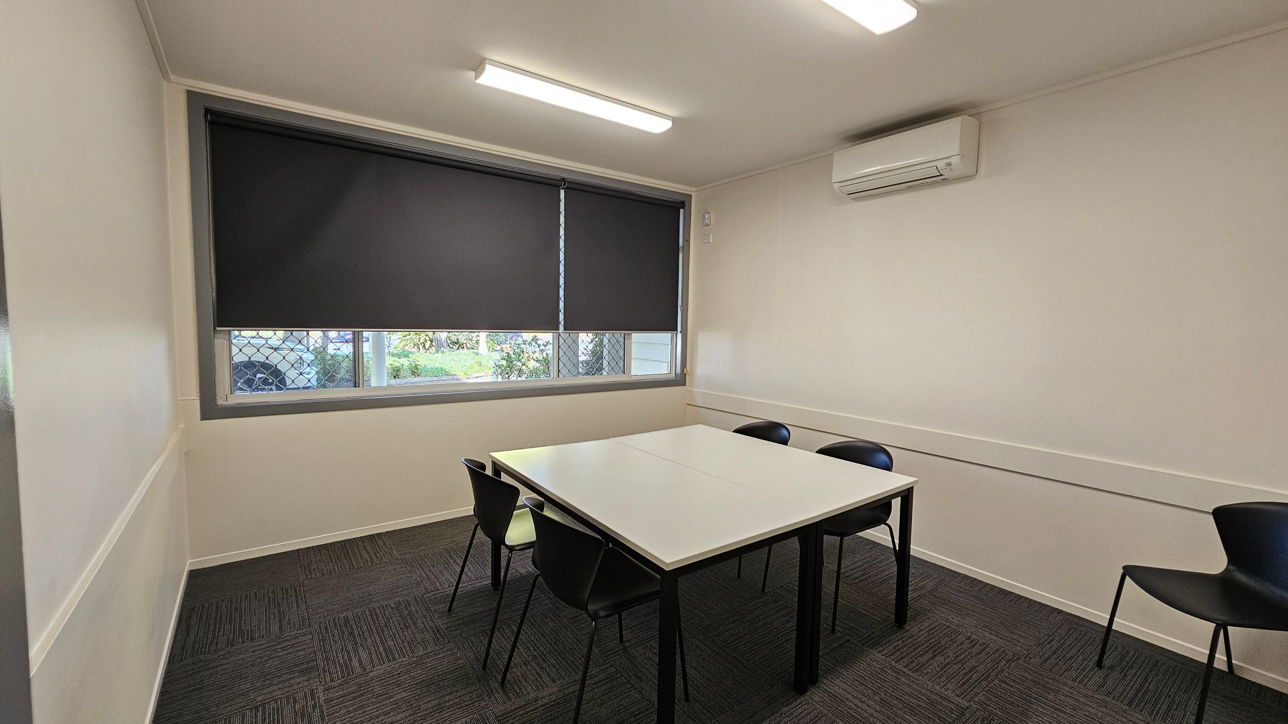 A small room with a square table in the centre, surrounded by four black chairs. The left wall features a large window with a dark roller blind partially down, allowing a glimpse of greenery outside. The right wall has a mounted air conditioning unit. The floor is covered with dark grey carpet tiles, and the ceiling has two fluorescent lights. The walls are light-coloured, creating a simple and functional space.