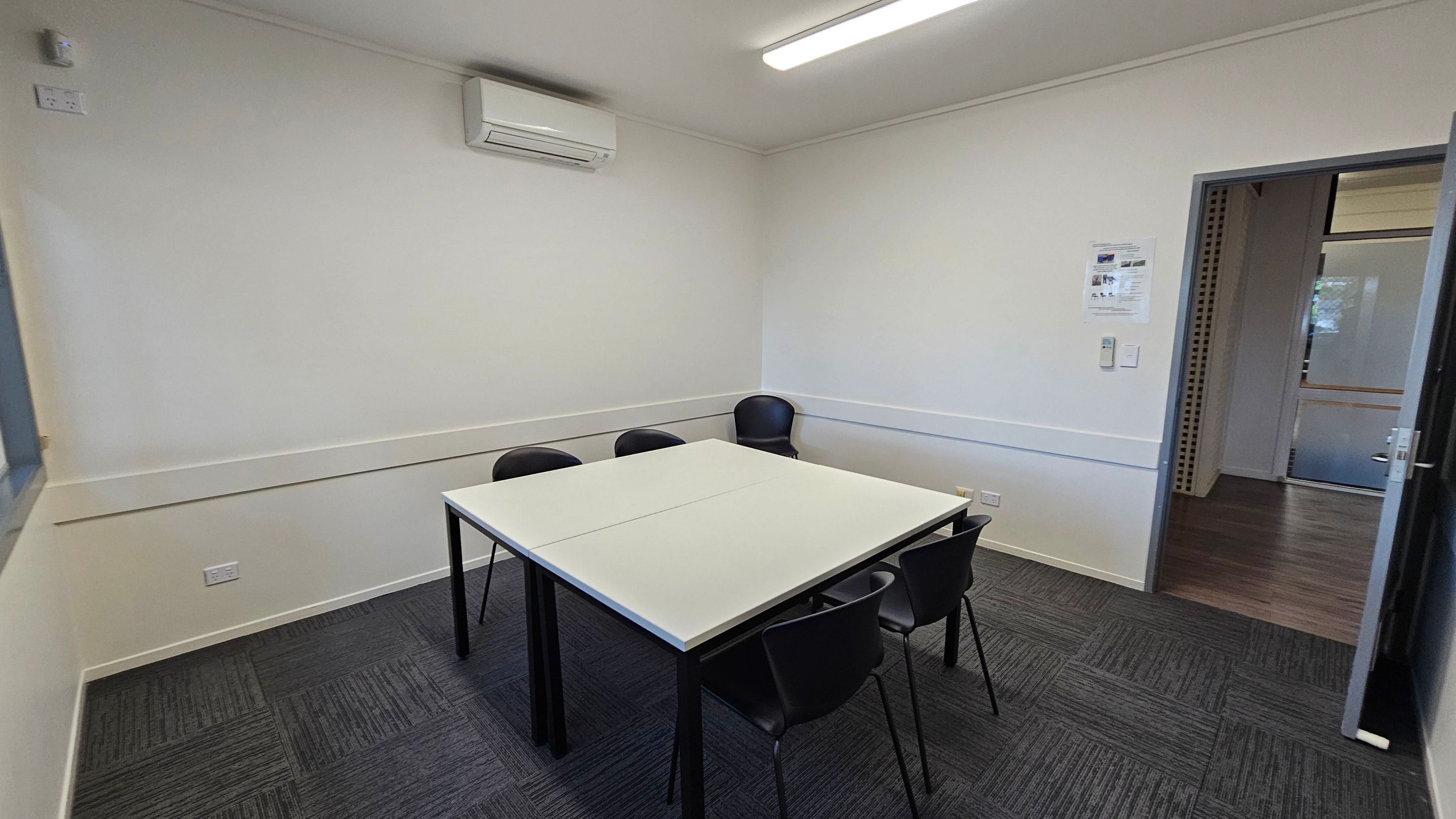 A small meeting room with a square table in the centre surrounded by four black chairs. The walls are plain white, and there's an air conditioning unit mounted on the left wall. The floor is covered with dark grey carpet tiles. On the right wall, there is a door leading to another room with wooden flooring. A fluorescent light fixture is on the ceiling, and a small notice is posted on the right wall near the door.