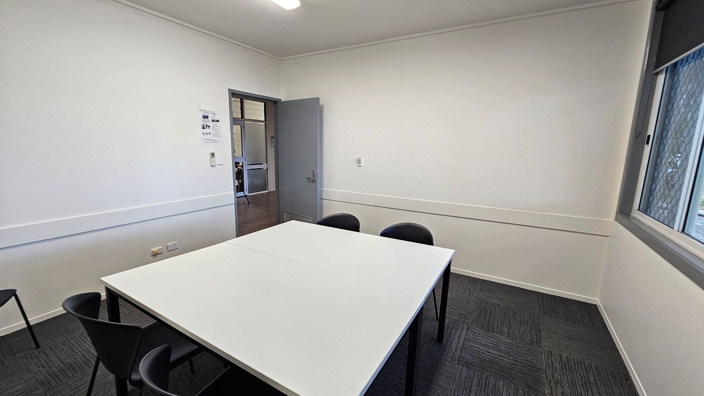 A small meeting room with a white table in the centre surrounded by four black chairs. The walls are plain white, with a door on the left leading to another room. A window with a mesh screen is on the right wall. The floor is covered with dark grey carpet tiles. A fluorescent light fixture is on the ceiling.