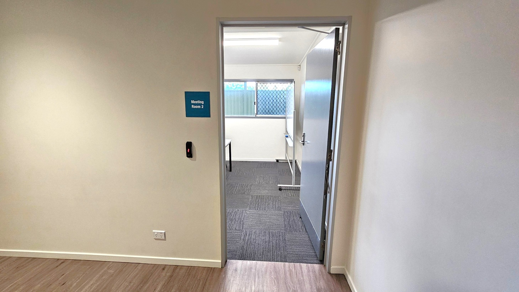 A doorway leads into a room labelled "Meeting Room 2" on a blue sign. The door is open, revealing a room with grey carpet tiles and a window with a mesh screen. A table is partially visible inside. The walls are light-coloured, and the floor outside the room is wooden. A small black device is mounted on the wall near the door, and there is an electrical outlet below it. The ceiling inside the room has a fluorescent light fixture.