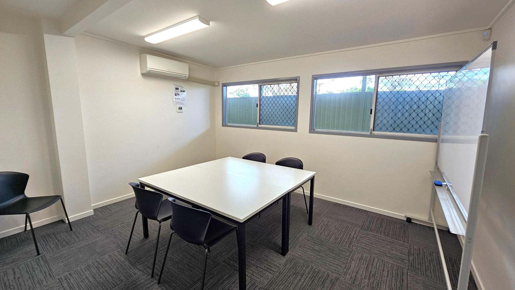 A small room with a white ceiling and fluorescent lights. On the left, there's a wall-mounted air conditioner above a couple of posters. Below, a black chair is placed against the wall. In the centre, a square table with a white top is surrounded by four black chairs. On the right, two windows with security grilles let in natural light. A whiteboard is positioned against the right wall. The floor is covered with dark grey carpet tiles.