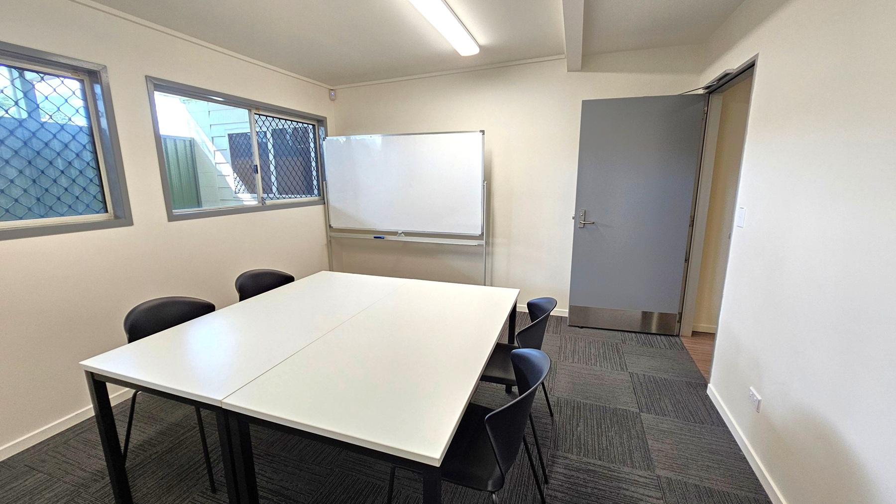 A small meeting room with a white rectangular table in the centre, surrounded by black chairs. On the right, a grey door is slightly open. The back wall features a whiteboard. On the left, two windows with security grilles allow natural light in. The floor is covered with dark grey carpet tiles, and a fluorescent light fixture is mounted on the ceiling.