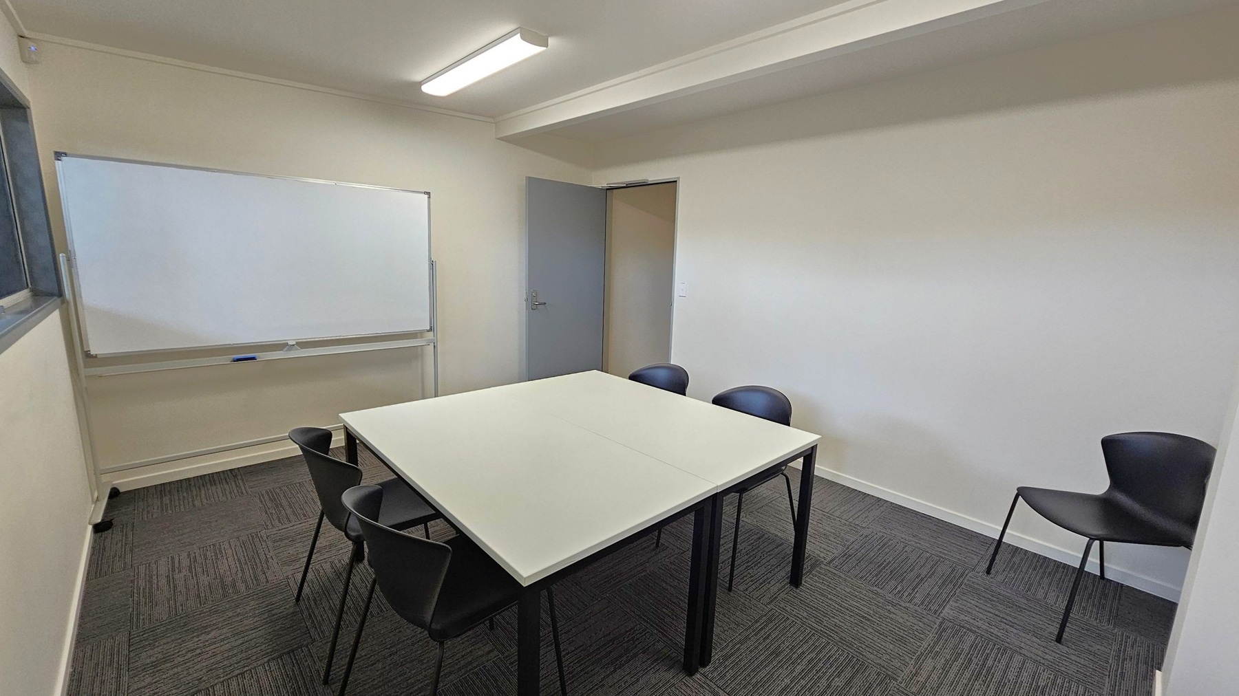 A small meeting room with a square table in the centre surrounded by five black chairs. On the left, a large whiteboard is mounted on the wall. The floor is covered with dark grey carpet tiles. A grey door is visible in the background, slightly ajar. The walls are light-coloured, and a fluorescent light fixture is on the ceiling.