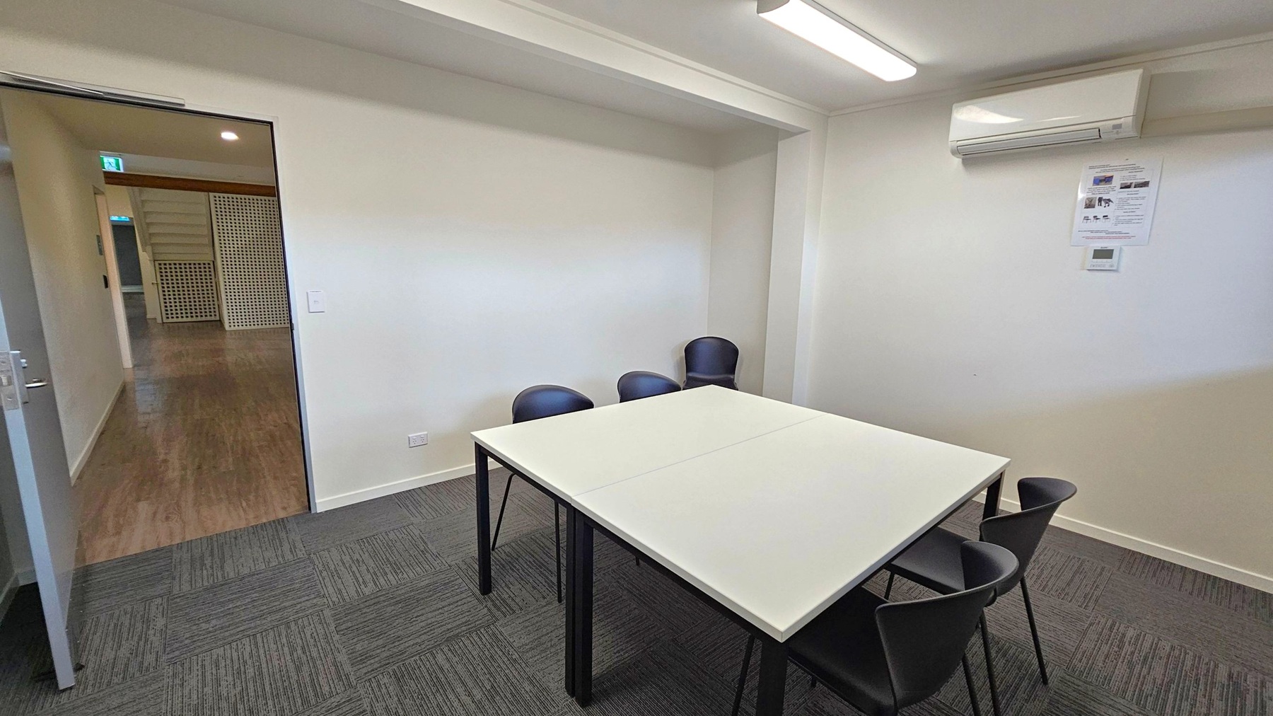 A small meeting room with a white table in the centre surrounded by four black chairs. The floor is covered with dark grey carpet tiles. On the right wall, there is an air conditioning unit and a small poster. The ceiling has a fluorescent light fixture. An open door on the left leads to a hallway with wooden flooring and a green lattice partition.