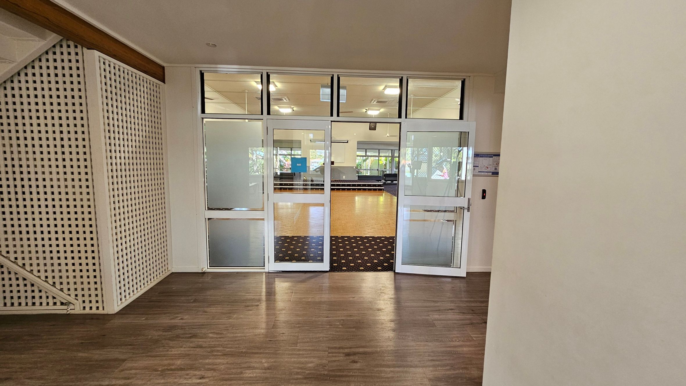 A hallway with wooden flooring leads to a set of glass double doors. The doors open into a room with a patterned carpet and wooden flooring. On the left, a white lattice panel is visible, possibly part of a staircase. The ceiling has recessed lighting, and the room beyond the doors is well-lit with large windows in the background. A small blue sign is visible on the left door.