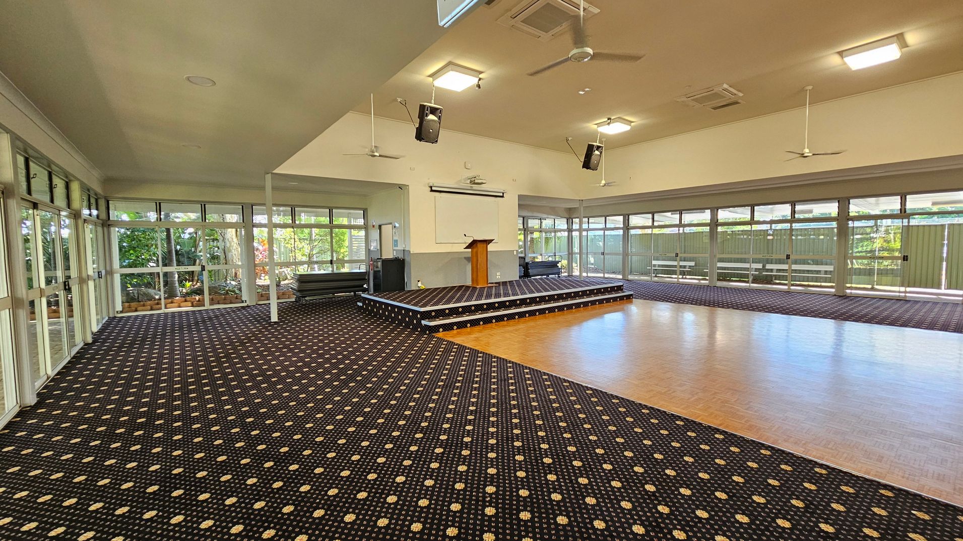 A spacious room with a patterned carpet on the left and a wooden floor on the right. In the centre, a raised stage with a wooden podium and a blank screen behind it. The room has large windows along the walls, allowing natural light to enter. Ceiling fans and speakers are mounted on the ceiling. The walls are light-coloured, and there are plants visible outside through the windows.