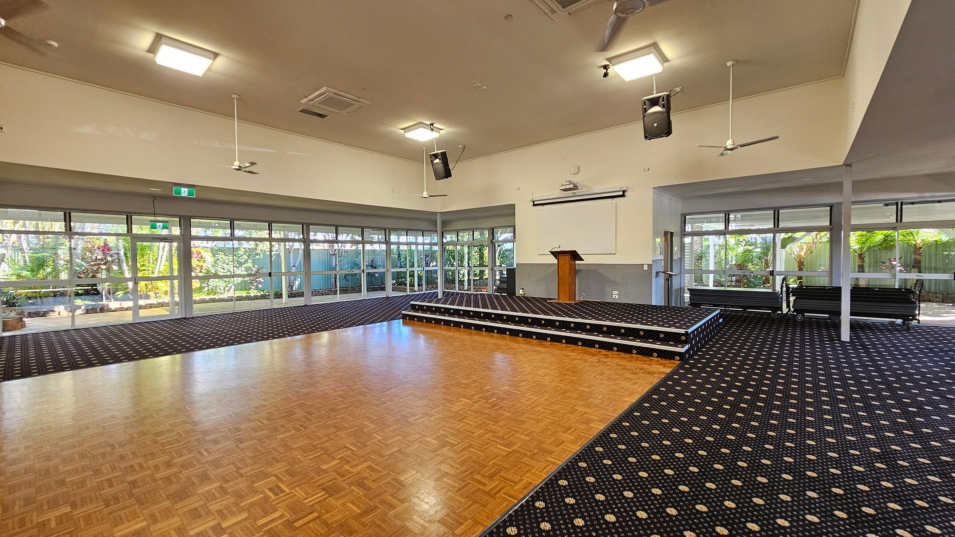 A spacious room with a parquet wooden floor in the centre and patterned carpet around the edges. On the right, there's a small stage with a lectern and a projection screen above it. The left side features large windows and glass doors, revealing a garden with lush greenery outside. The ceiling has several square lights, ceiling fans, and speakers. The walls are light-coloured, and there are stacked chairs on the right side near the stage.