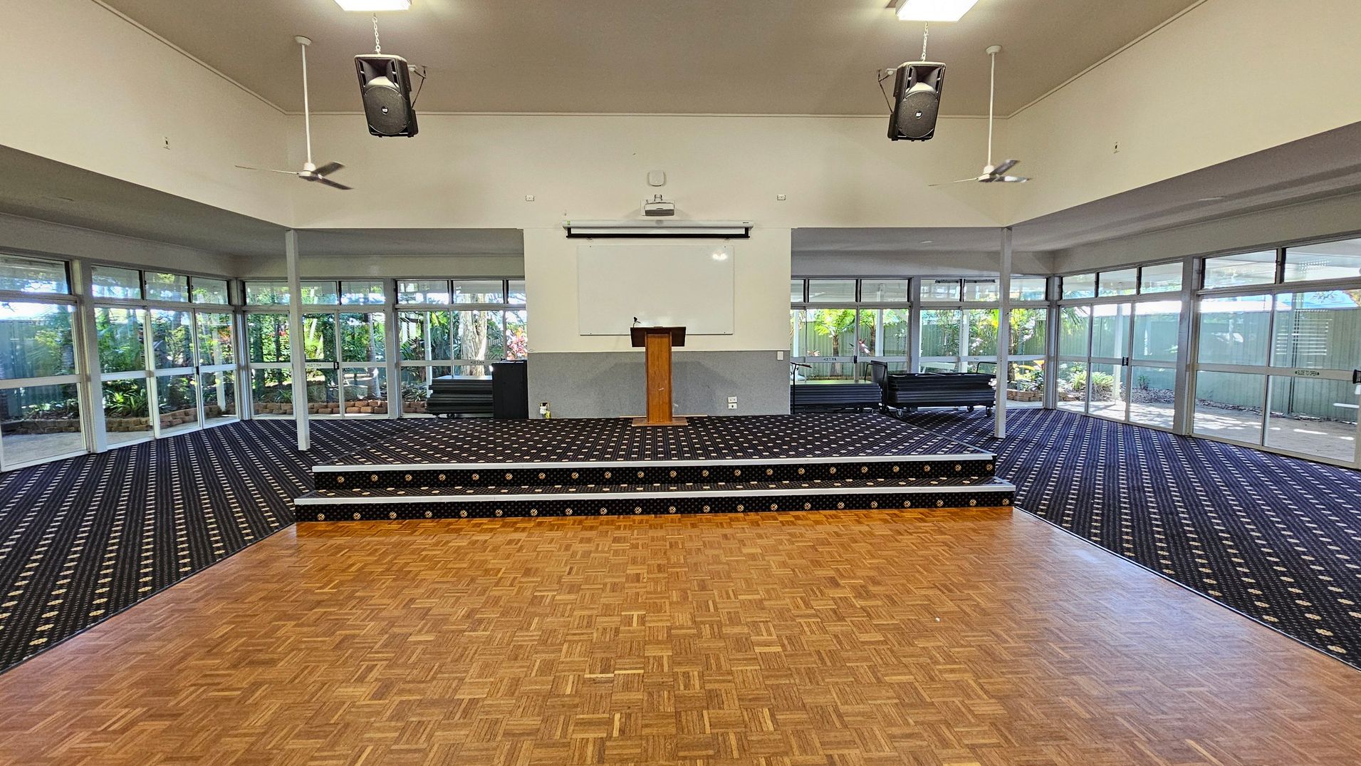 A spacious room with a parquet wooden floor in the foreground and a raised platform in the middle. The platform is carpeted with a dark patterned design and features a wooden lectern. Behind the platform is a whiteboard or screen. Large windows line the left and right walls, allowing natural light to illuminate the space. Ceiling fans and speakers are suspended from the ceiling, which is plain and light-coloured. The background shows greenery visible through the windows.