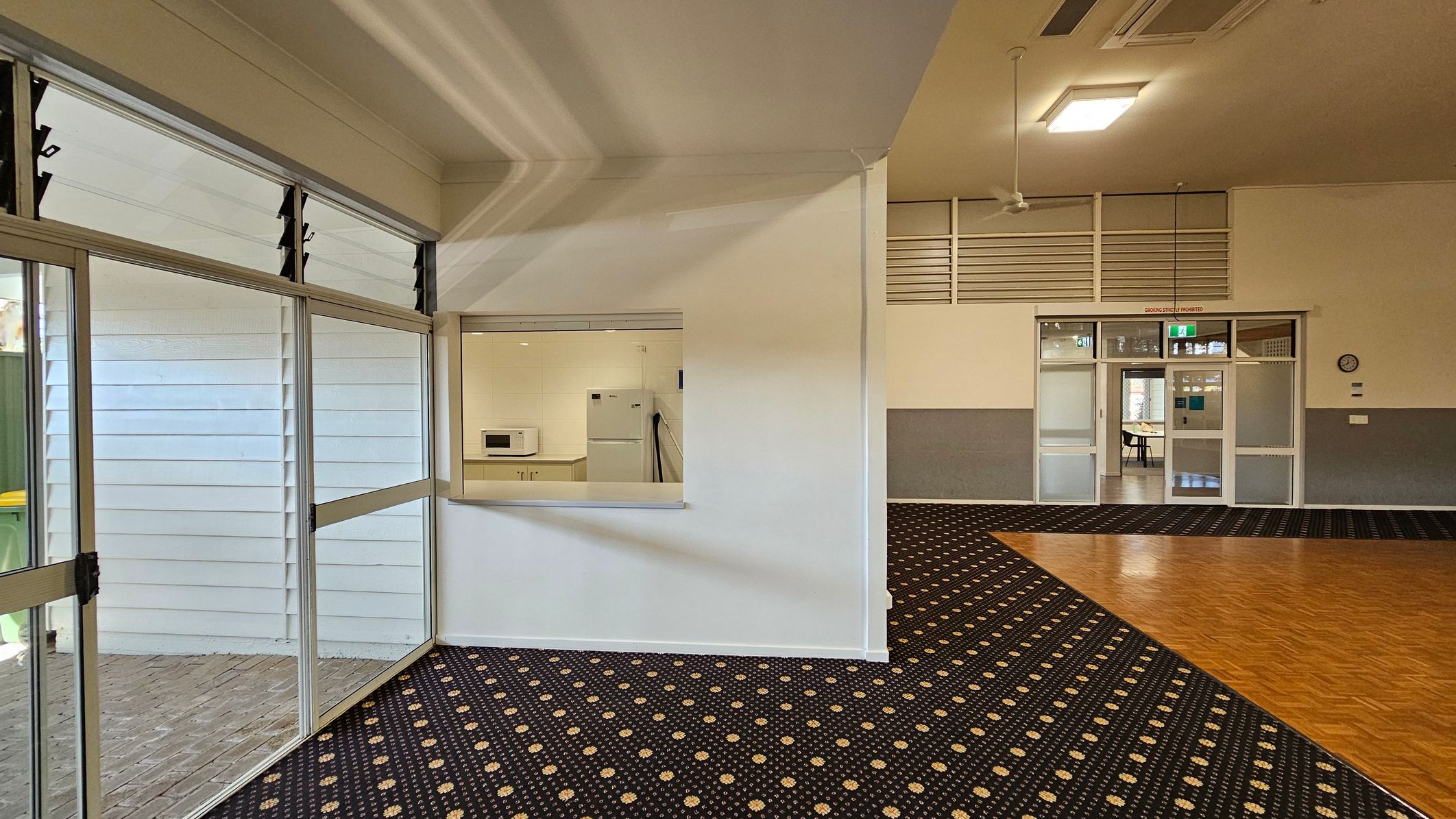 On the left, there are large glass windows with louvres, revealing an outdoor area with a yellow bin. In the middle, a small serving hatch opens into a kitchen area with a microwave and a fridge. On the right, a doorway with glass panels leads to another room, with a clock on the wall nearby. The floor is a mix of patterned carpet and wood, and the ceiling has fluorescent lights and a ceiling fan.