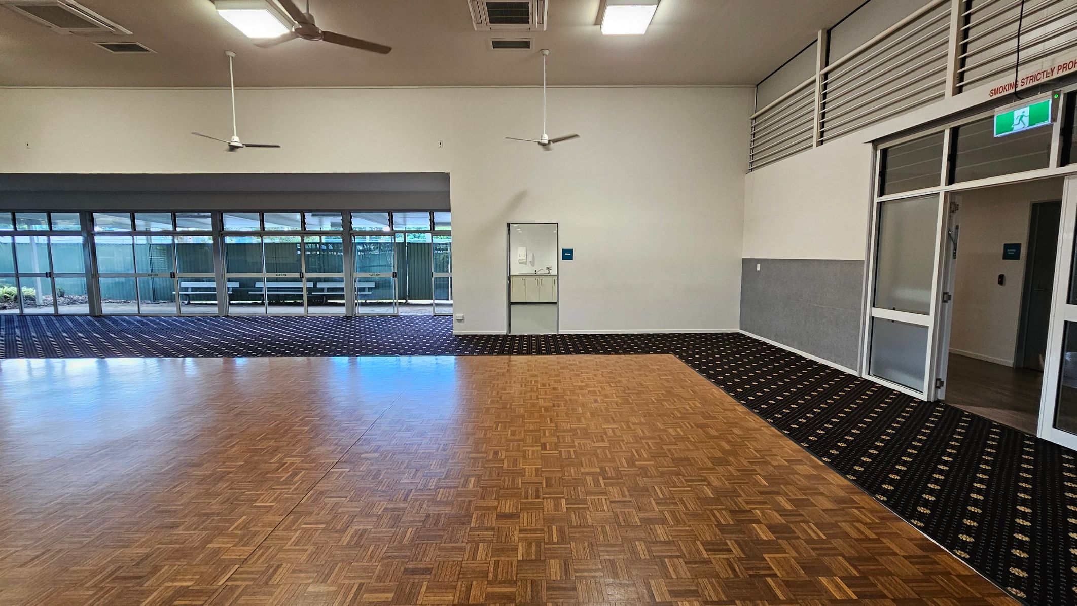 A spacious room with a parquet wooden floor in the foreground and patterned carpet in the background. On the left, large windows with glass doors open to an outdoor area with a bench. In the centre, a small door leads to a room with a sink. On the right, a glass door with an exit sign above it. The ceiling is equipped with ceiling fans and fluorescent lights. The walls are light-coloured with a grey panel halfway up.