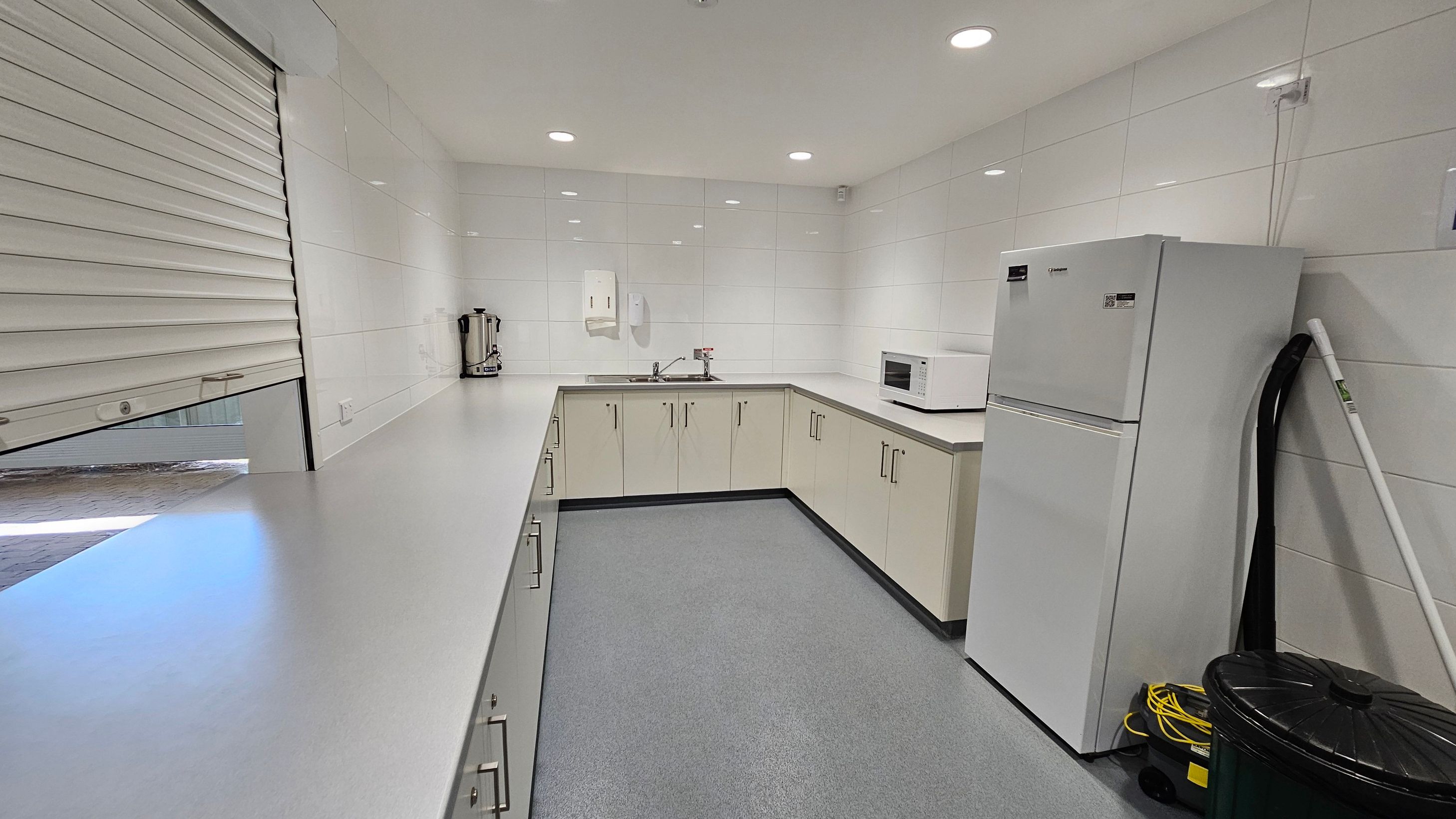 A clean, modern kitchen with white tiled walls and a grey floor. On the left, a long countertop with a closed roller shutter above it. In the middle, a corner sink with a paper towel dispenser on the wall. To the right, a white microwave sits on the counter, and a large white refrigerator stands next to it. A mop and bucket are placed beside the fridge. The ceiling has recessed lighting.