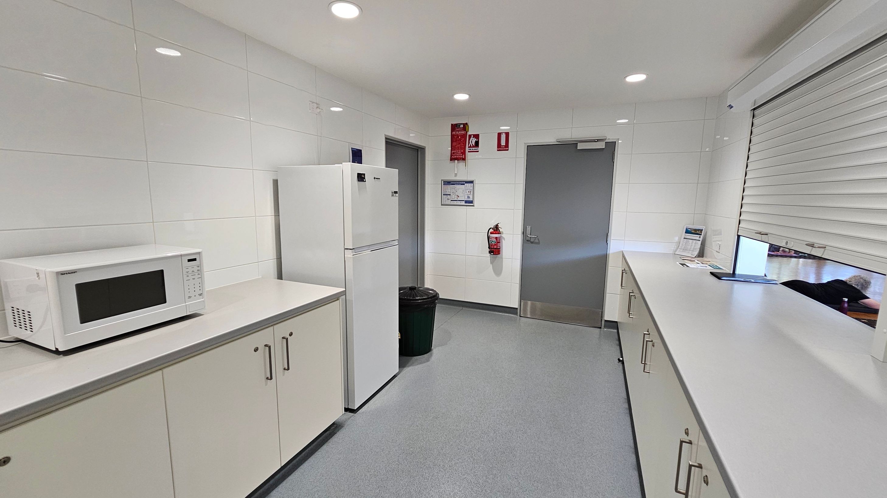 A clean, modern kitchen with white tiled walls and a grey floor. On the left, a white microwave sits on a countertop above light-coloured cabinets. Next to it is a tall white refrigerator. In the centre, a grey door is flanked by a fire extinguisher and a waste bin. On the right, a long countertop runs beneath a partially open shutter window, with some papers and a tablet on the surface. The ceiling is white with recessed lighting.