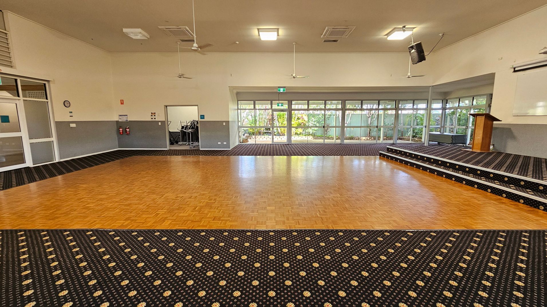 A spacious room with a parquet wooden floor in the centre, surrounded by patterned carpet. On the right, there is a raised stage with a wooden lectern. Large windows line the back wall, offering a view of greenery outside. On the left, there are doors and a small storage area with stacked chairs. The ceiling is equipped with ceiling fans and lights.