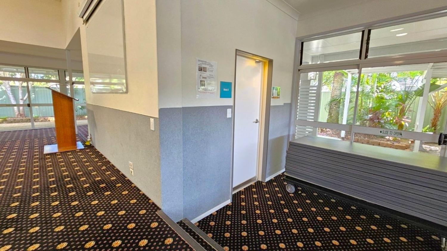 A room with a patterned carpet featuring yellow and black designs. On the left, there's a wooden podium near large windows with a view of trees and a fence outside. In the centre, a doorway labelled "Store 2" is visible, with some notices on the wall. On the right, a stack of tables on wheels is positioned in front of more windows, which have a "slide to open" sign. The ceiling is plain and light-coloured.