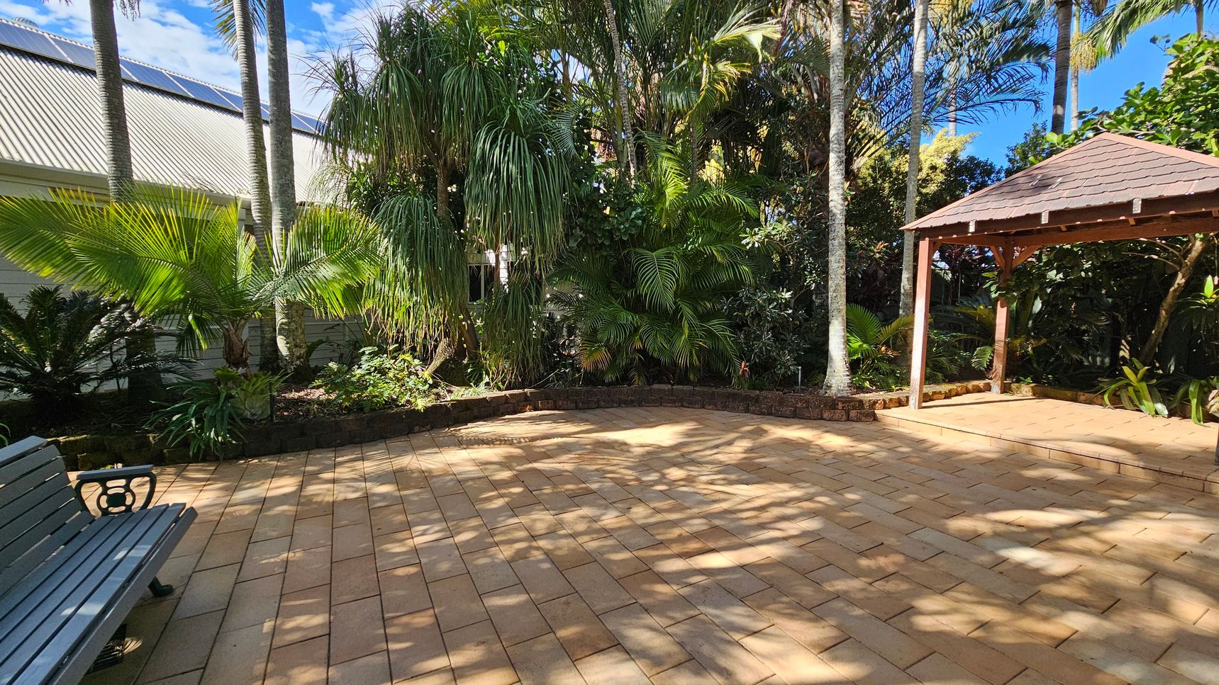A paved outdoor area with a wooden bench on the left. In the centre, there are various tropical plants and trees, including palm trees, creating a lush backdrop. On the right, a wooden gazebo with a shingled roof stands on the paved surface. The ground is covered with light brown paving stones, and the area is partially shaded by the surrounding foliage.