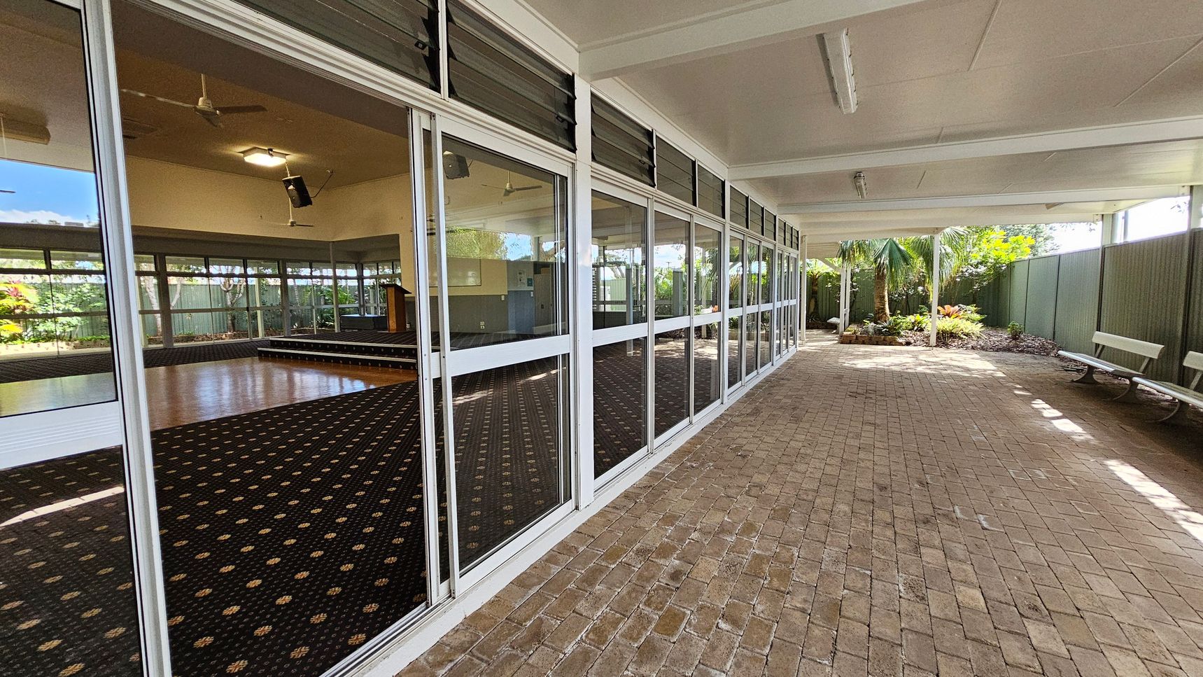 A covered outdoor area with a brick floor and a ceiling with fluorescent lights. On the left, there are large glass windows and doors revealing an interior space with a patterned carpet, a wooden stage, and ceiling fans. The right side features a green fence and a garden with palm trees and plants. Two white benches are positioned along the fence.