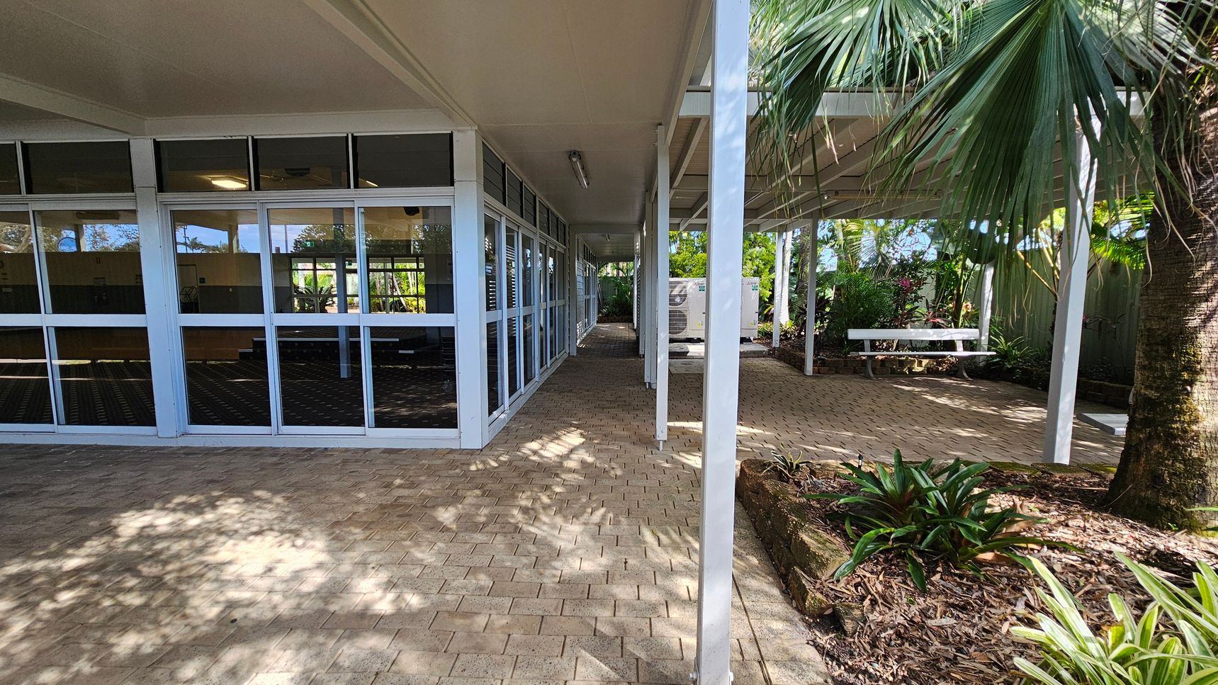 A covered outdoor walkway with a brick floor. On the left, there is a building with large glass windows reflecting the surroundings. The right side features a garden area with a variety of plants, including a large palm tree. A white bench is situated under the covered area, surrounded by greenery. The ceiling is flat and white, with a fluorescent light fixture.