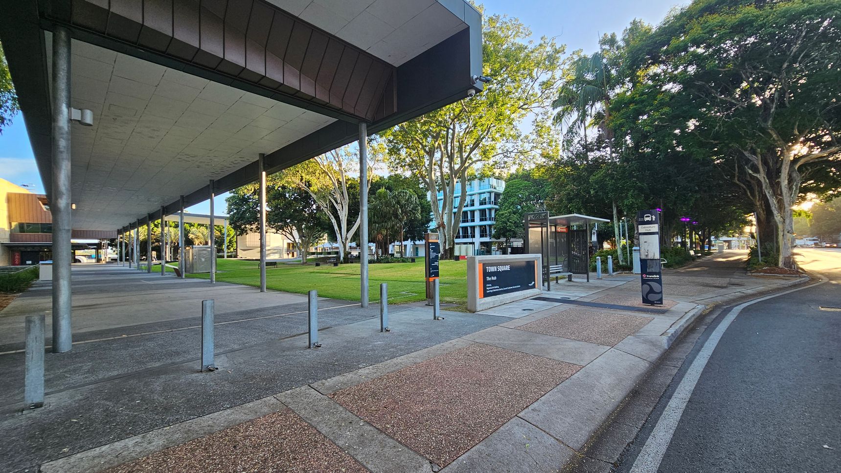 A modern urban area with a wide walkway on the left, covered by a large overhanging roof supported by metal poles. The ground is a mix of concrete and textured paving. In the centre, there's a grassy area with trees and a sign reading "Town Square." On the right, a bus stop with a shelter and a signpost is visible. The background features a multi-storey building partially obscured by trees. The scene is well-lit, suggesting early morning or late afternoon.