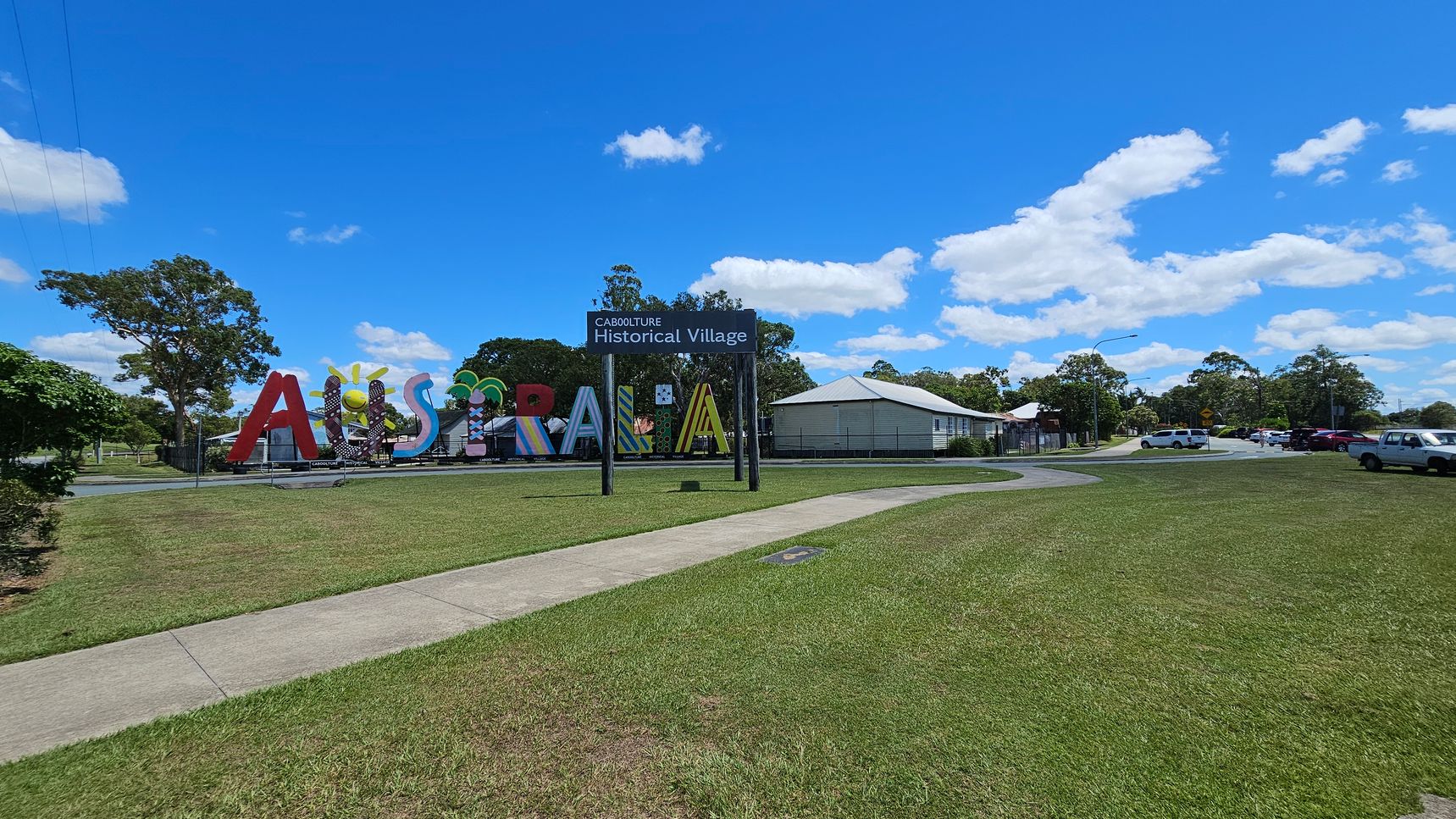Large colourful letters spelling "Australia" are on the left, with a sign above reading "Caboolture Historical Village". A white building with a sloped roof is in the centre-right. Several cars are parked on the right side near a road. The foreground features a grassy area with a concrete path, while trees are scattered in the background under a bright blue sky with scattered clouds.