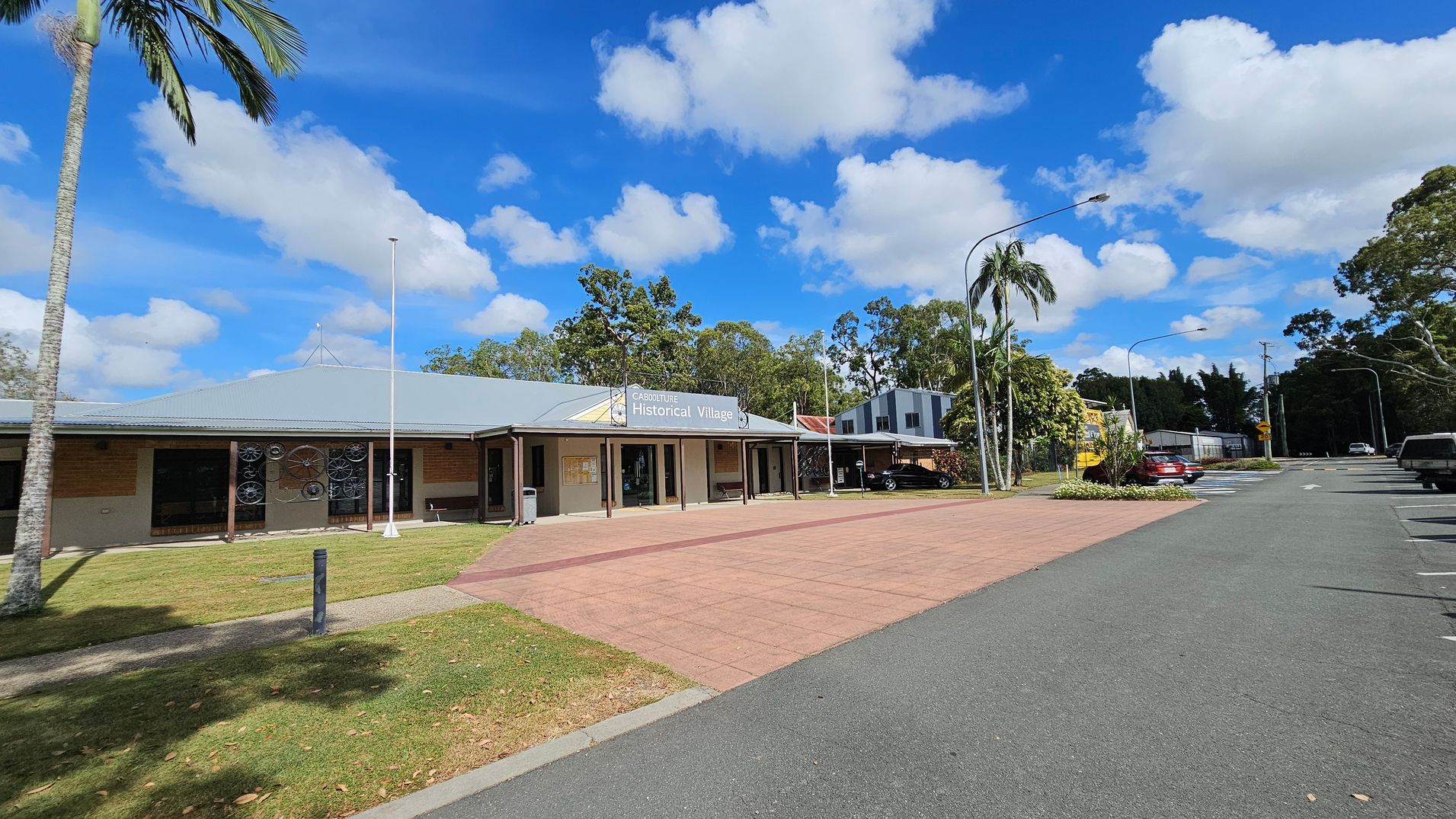 A single-storey building with a grey roof and a sign reading "Caboolture Historical Village" is in the centre. The building has a veranda with several poles and decorative wheels on the wall. To the right, a car park with several vehicles and street lamps is visible. A palm tree and other greenery are on the left, with a paved walkway and grass in the foreground. The background features more trees and a partly cloudy sky.