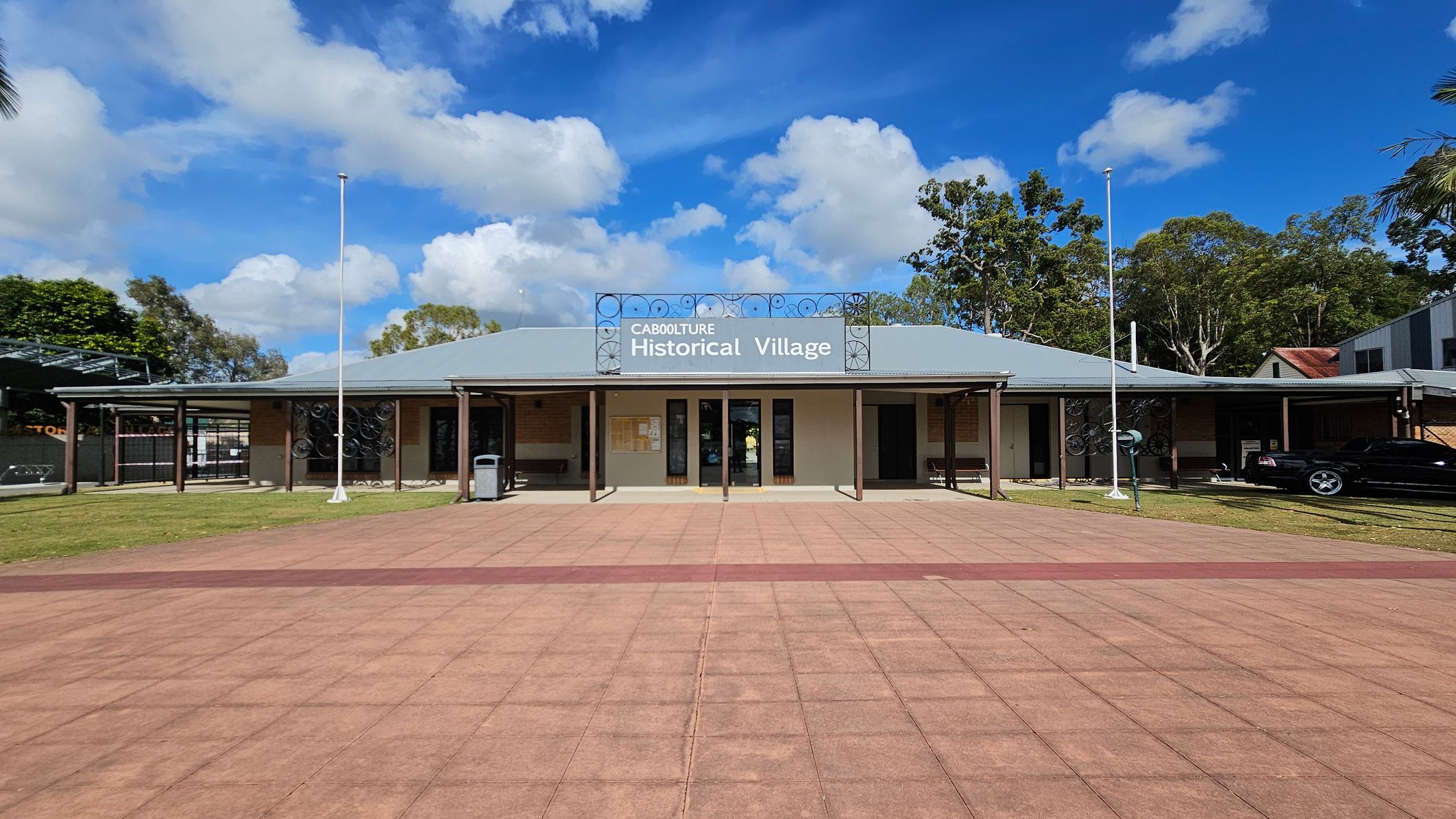 A single-storey building with a grey roof and a sign reading "Caboolture Historical Village" at the centre. The building has a covered walkway supported by poles, with a bin on the left. Two flagpoles stand in front, one on each side. The foreground features a paved area with a reddish-brown pattern. Trees and a parked car are visible on the right, while more trees and a structure are on the left.
