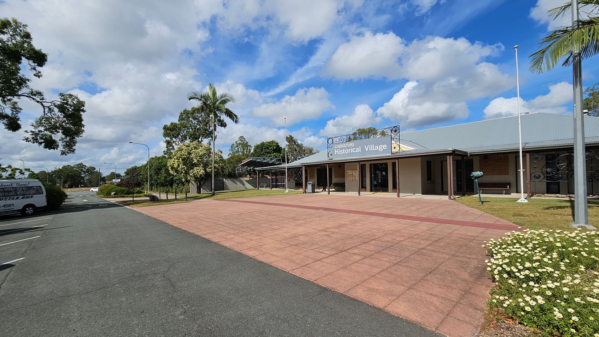 A paved area leads to a building on the right with a sign reading "Caboolture Historical Village." The building has a grey roof and a veranda. A flagpole stands nearby, and a small garden with white flowers is in the foreground. On the left, a road with parked vehicles, including a van, extends into the distance. Trees and greenery are visible throughout the scene.