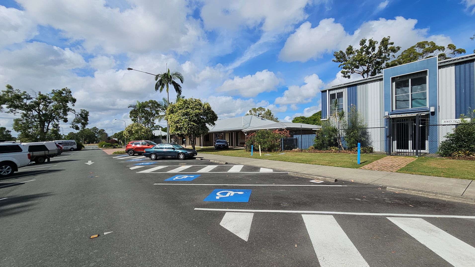 A street scene with a row of parked cars on the left, including a white pickup and a red SUV. On the right, there are designated disabled parking spaces marked in blue. A building with blue and grey panels is visible, featuring large windows and a gated entrance. Trees and shrubs are scattered throughout, with a palm tree near the centre. The road has a white arrow indicating direction, and a brick pathway leads to the building. The pavement is bordered by grass.