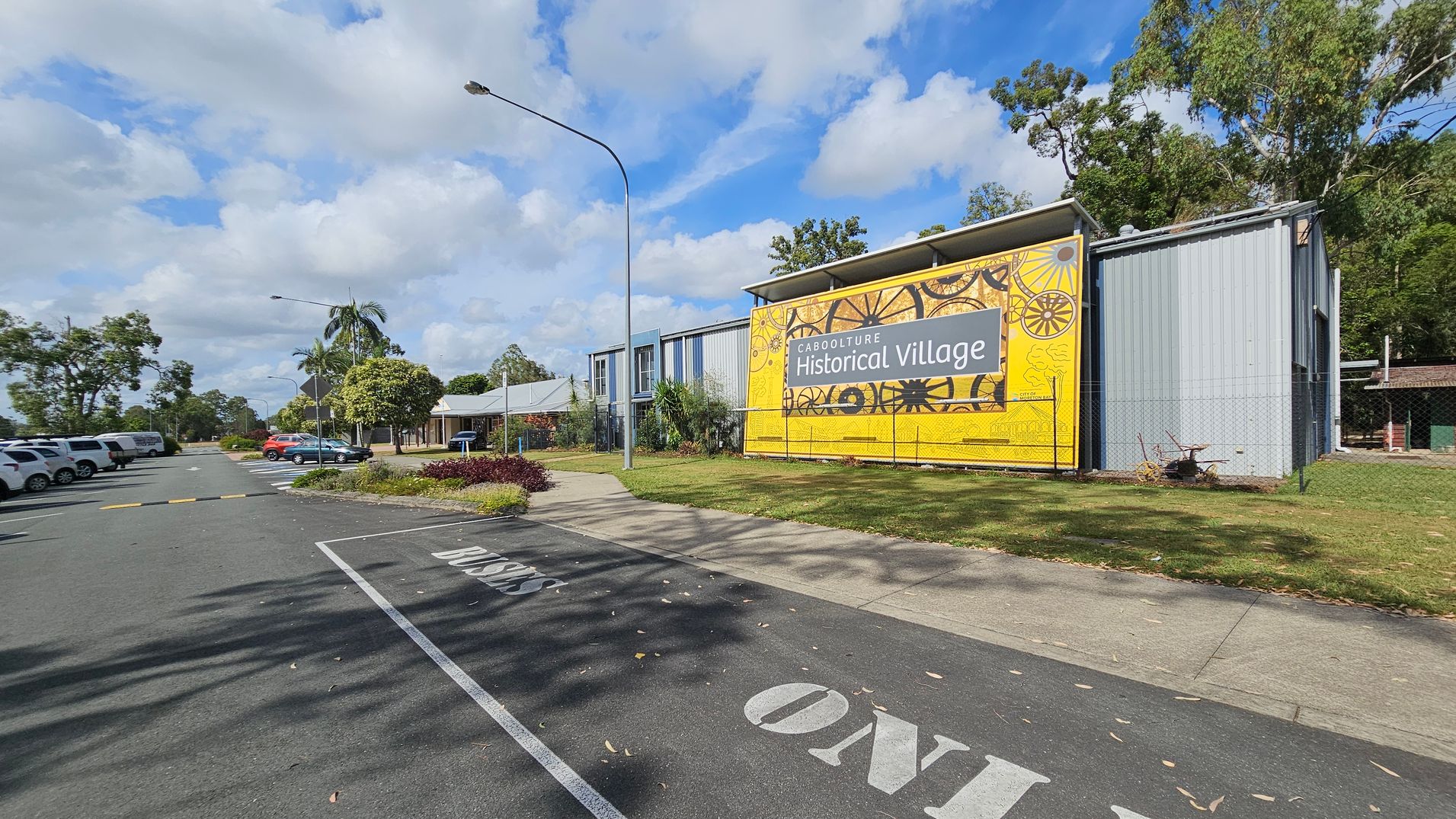 A large yellow sign on the right reads "Caboolture Historical Village" with decorative wheel designs. The building is grey with a metal exterior. A paved path runs alongside, bordered by grass. On the left, a row of parked cars lines the street, with a "Buses Only" lane marked on the road. Trees and shrubs are scattered throughout, and a tall streetlamp stands near the sign. The sky is partly cloudy.
