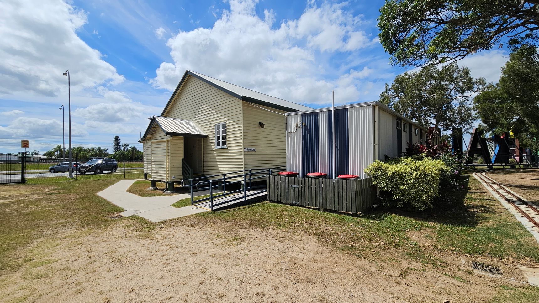 A small, light-coloured building with a metal roof and a ramp is on the right. The building has a sign indicating toilets. Next to it is a corrugated metal structure with red-lidded bins in front. A path leads to the building, surrounded by grass and dirt. In the background, there are trees and a large sign with letters. On the left, a road with cars and streetlights is visible, separated by a fence.
