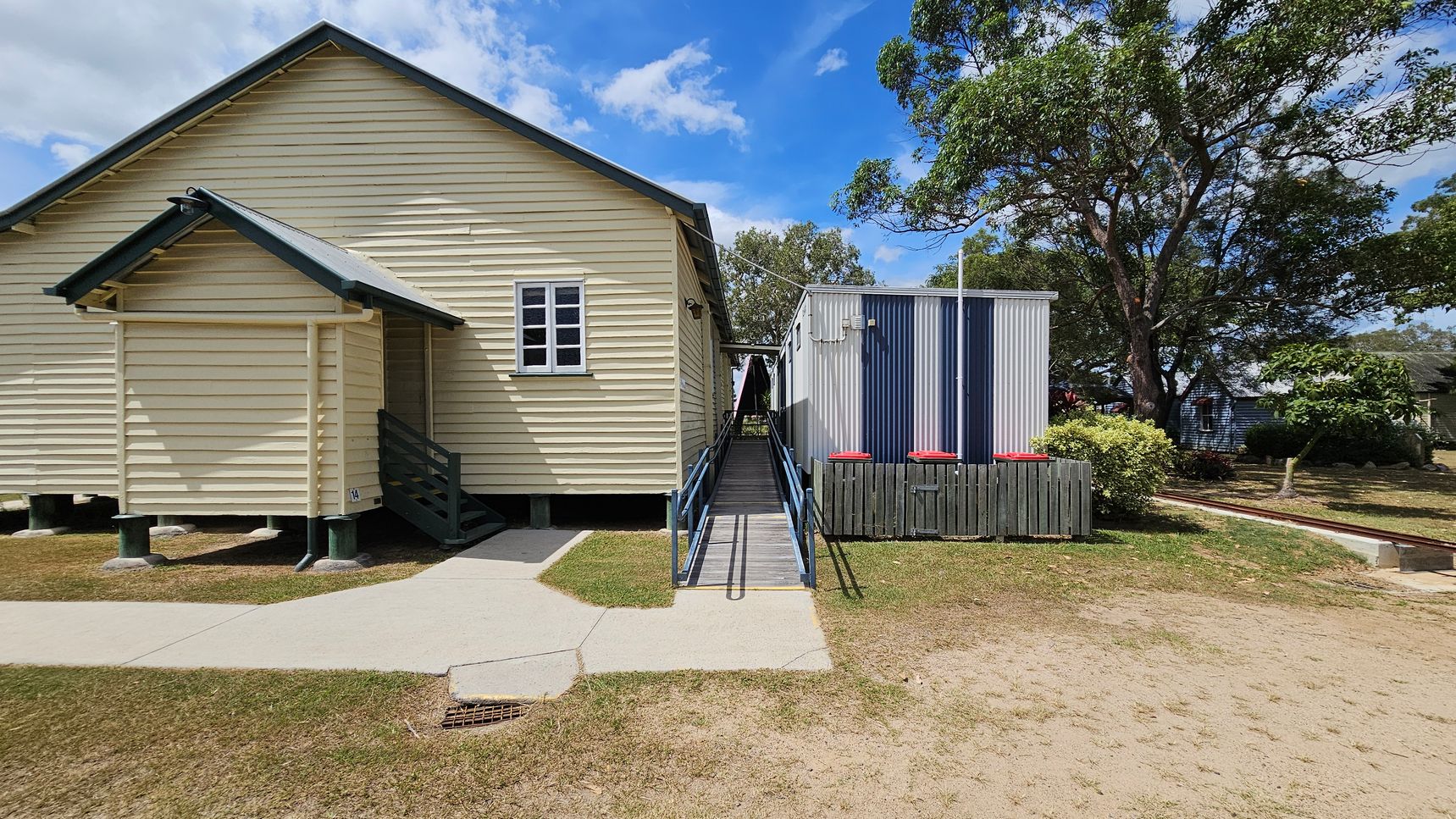 A light yellow wooden building with a small porch and steps is on the left. A metal ramp with blue railings leads to a grey corrugated metal structure on the right. The ground is a mix of grass and dirt, with a concrete path. In the background, there are trees and another building partially visible.