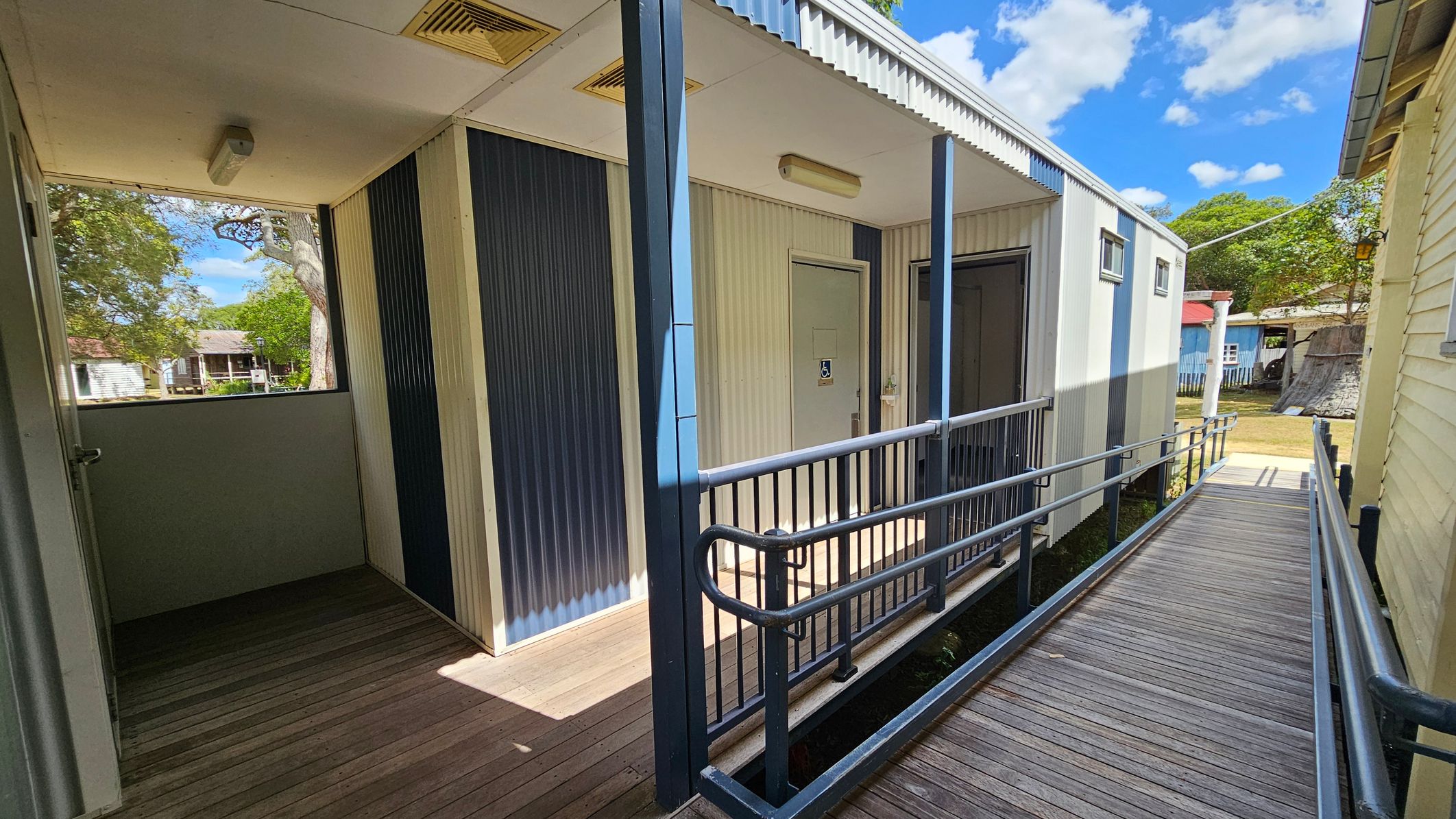 A wooden ramp with metal railings leads to a building with corrugated metal walls. The building has a door with a disability access sign. On the left, there's a small covered area with a window and a light fixture on the ceiling. In the background, there are trees and other small buildings. The sky is partly cloudy.