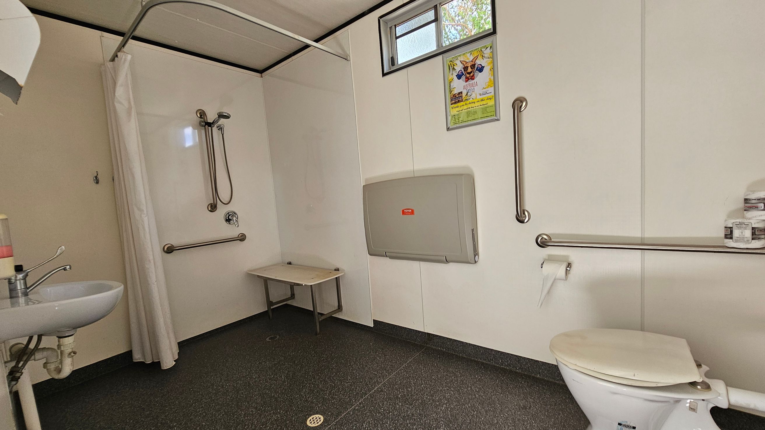 A bathroom with accessibility features. On the left, a small sink with a soap dispenser and a shower area with a curtain, handheld showerhead, and grab bars. In the centre, a foldable baby changing station is mounted on the wall beneath a small window. On the right, a toilet with grab bars and a toilet roll holder. A shelf above the toilet holds several small containers. The floor is covered with non-slip green tiles, and the ceiling is plain white.