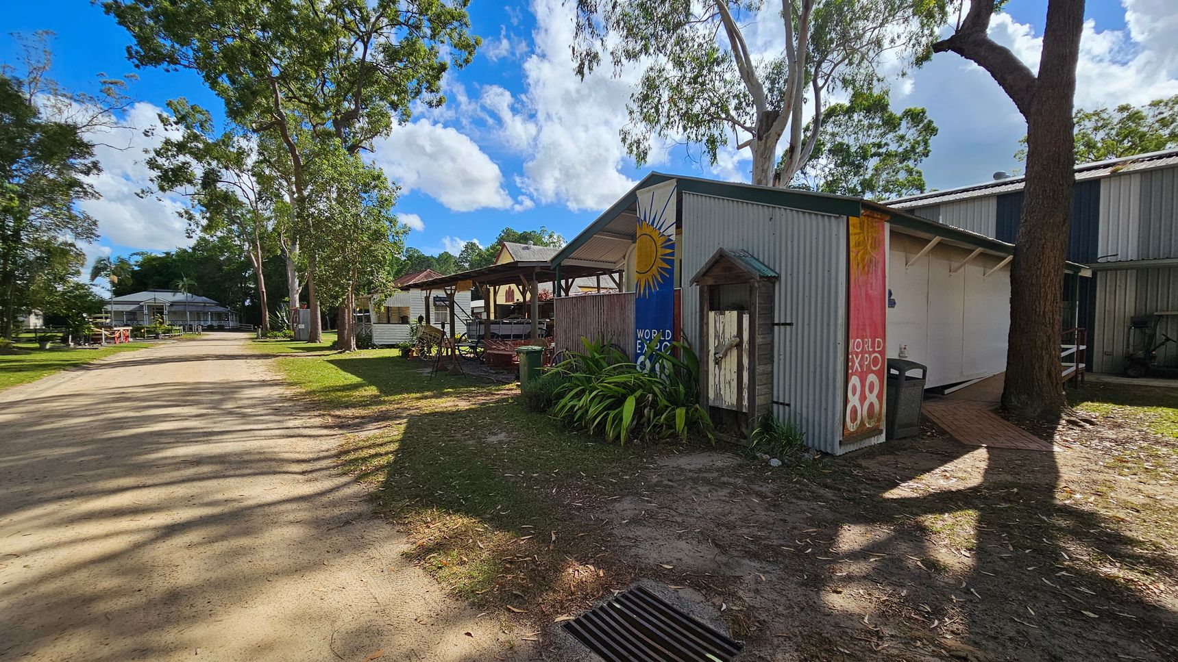 A dirt path runs through a grassy area with trees on both sides. On the right, there is a corrugated metal building with a colourful mural featuring a sun and the text "World Expo 88". Plants are in front of the building, and a tree casts a shadow on the ground. In the background, more buildings and trees are visible, with a clear blue sky and scattered clouds above.