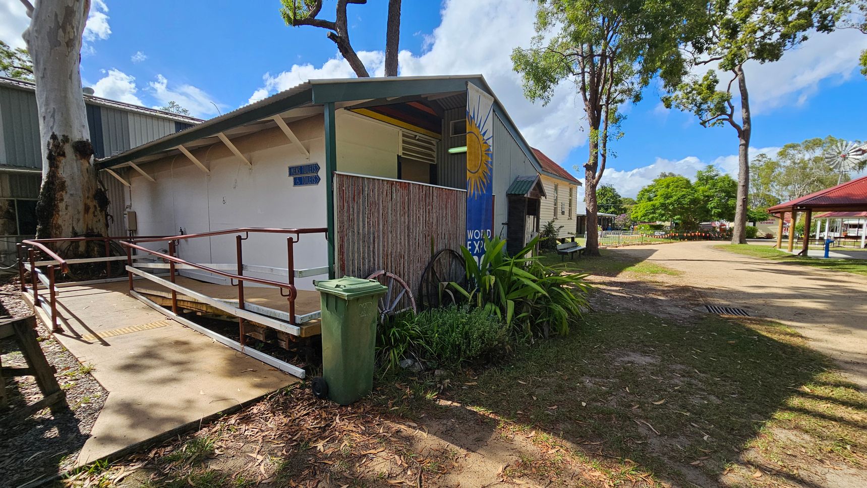 A small building with a ramp and handrails on the left, featuring a sign for toilets. The building has a mural with a sun design. A large tree is adjacent to the structure. In the background, there are more trees, a grassy area, and a red-roofed pavilion. A green wheelie bin and some plants are near the ramp. The ground is a mix of grass and dirt, with scattered leaves.