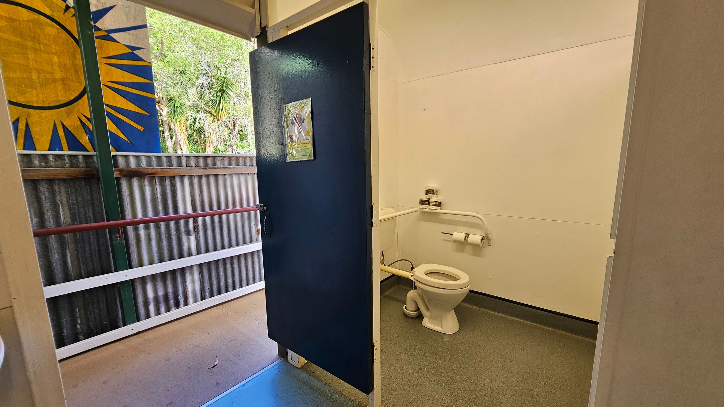 A public toilet with a partially open blue door. Inside, a white toilet with a grab rail and toilet paper holder is visible against a plain wall. The floor is grey. Outside, a corrugated metal wall with a colourful mural featuring a yellow sunburst on a blue background. Green foliage is visible in the background. The floor outside is a light brown.
