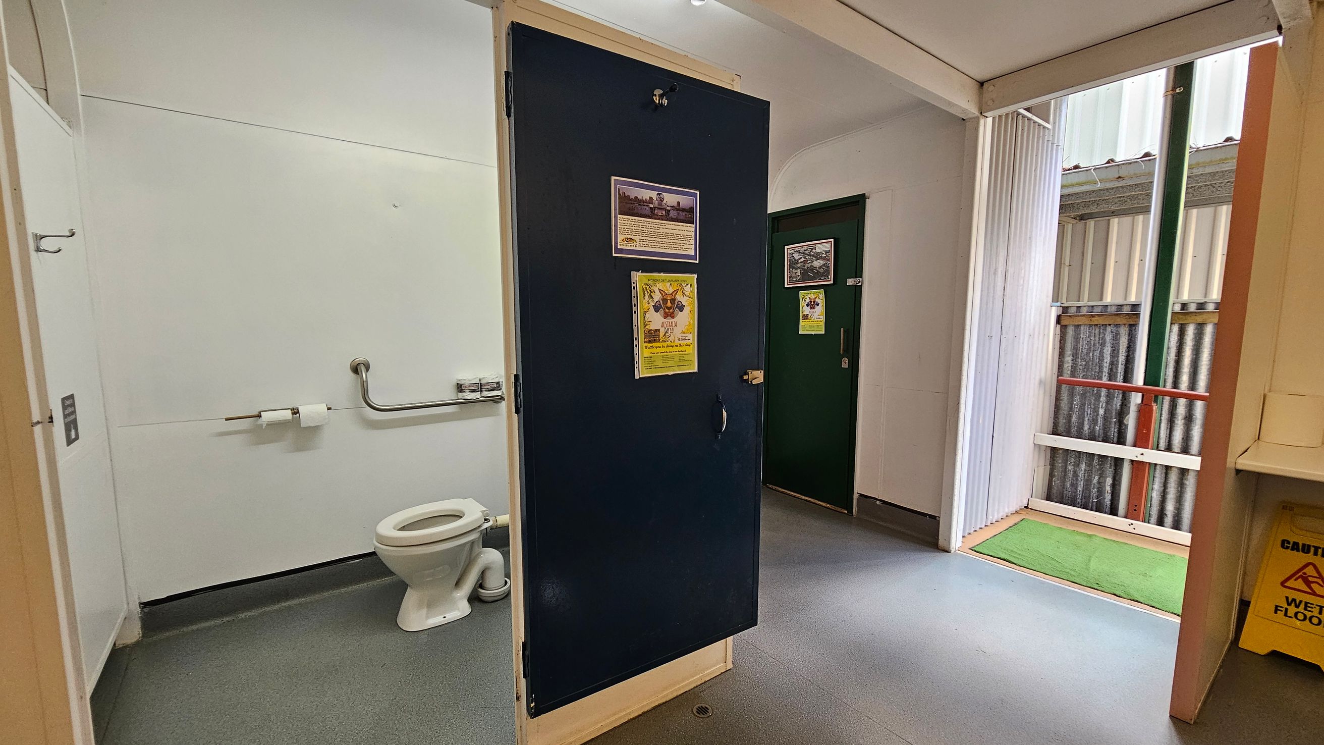 A public toilet with a white toilet and grab bar on the left wall. The floor is grey with a drain near the centre. A dark door with posters is partially open in the middle. On the right, there's an open entrance with corrugated metal walls and a green mat on the floor. A yellow "Caution: Wet Floor" sign is visible near a white sink. The ceiling is plain and white.