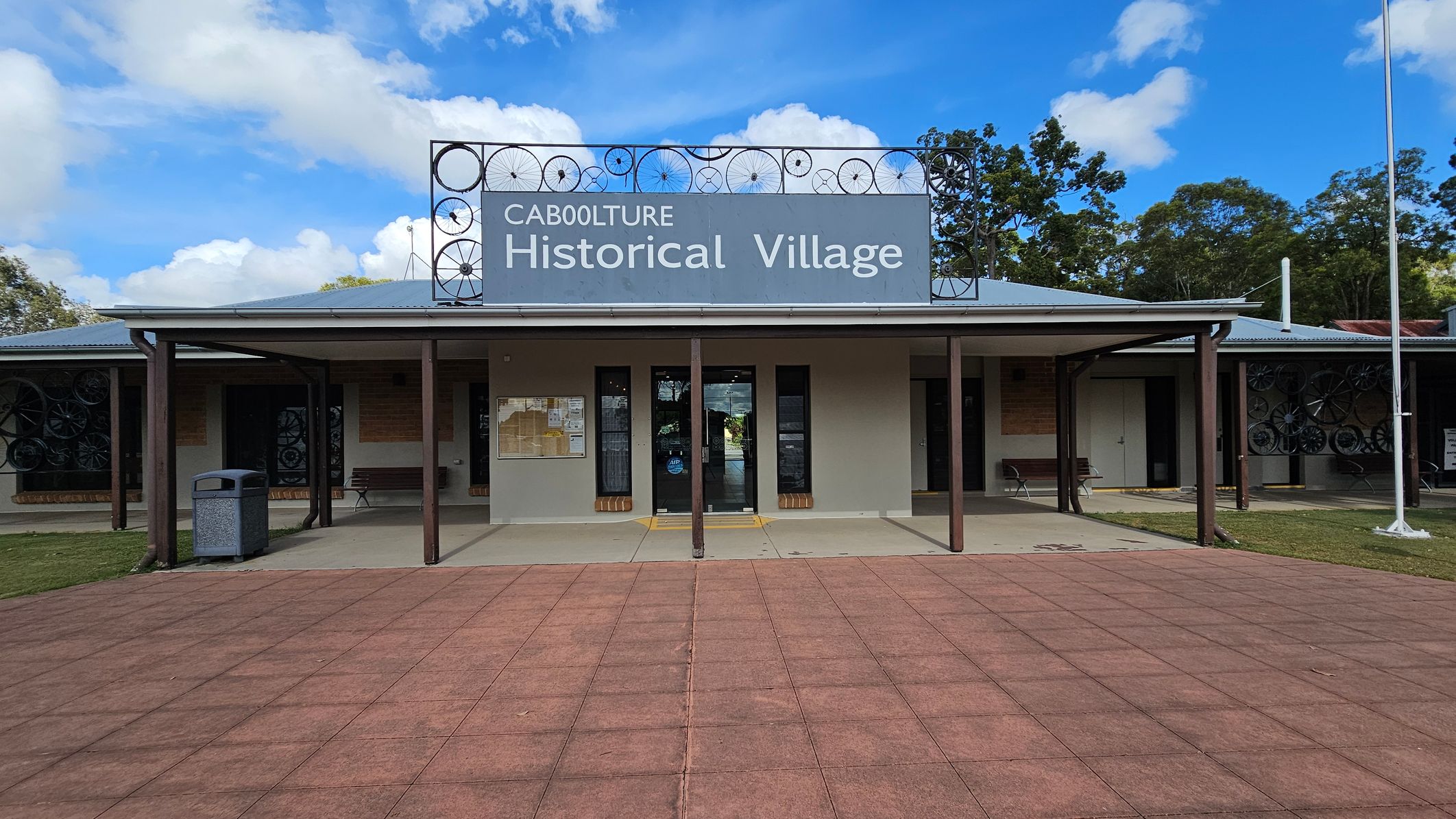 A building with a sign reading "Caboolture Historical Village" is centred in the image. The structure has a grey roof and a covered entrance supported by brown posts. Decorative metalwork featuring wheel designs is visible above the sign. The foreground consists of a red-tiled pathway leading to the entrance. On the left, there is a rubbish bin and a bench, while trees are visible in the background. The right side shows more benches and a flagpole.