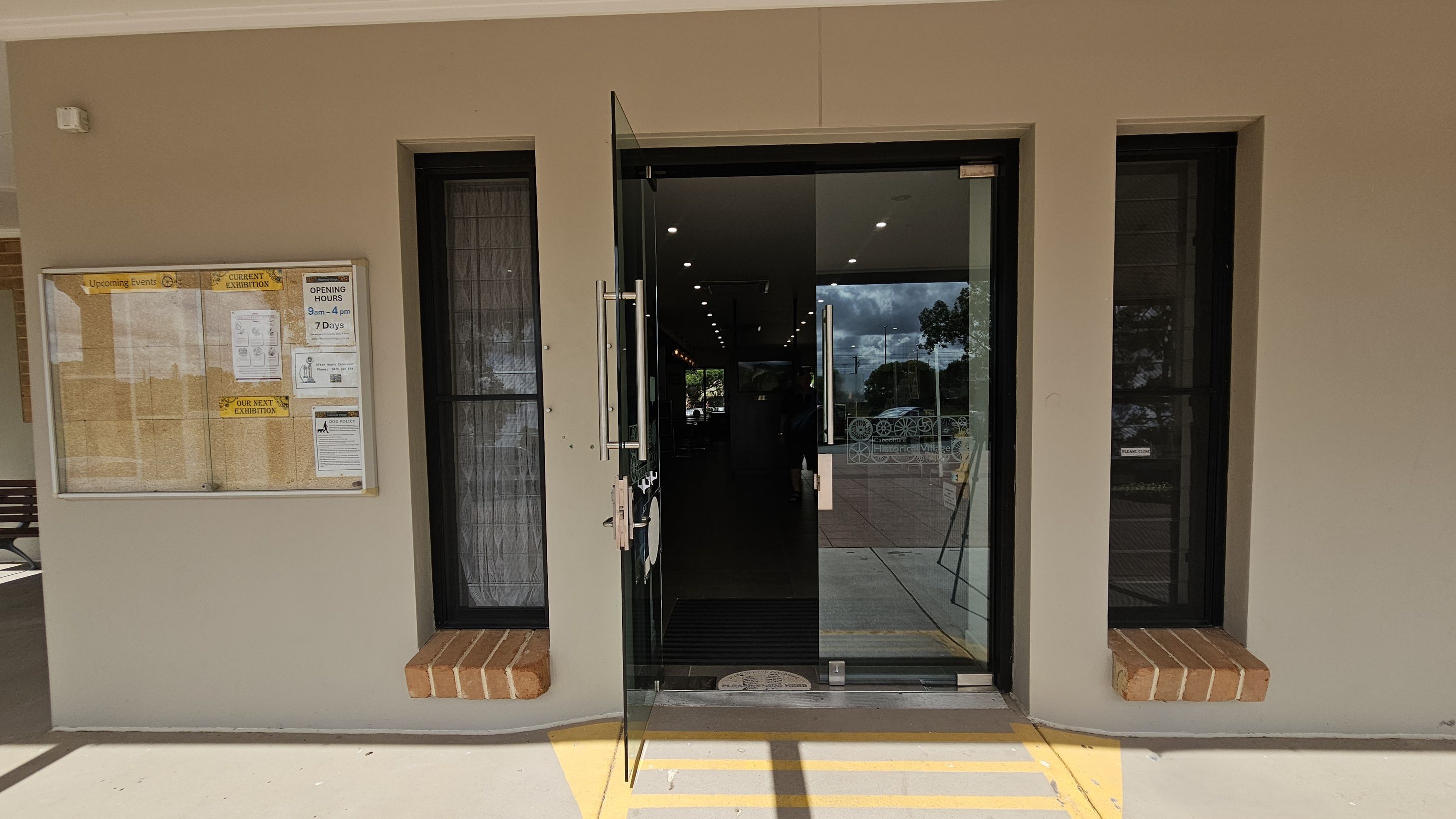 A glass door is centrally positioned, slightly open, with metal handles. On either side, there are narrow windows with black frames and brick sills. To the left, a notice board displays various papers and announcements, including opening hours. The floor is light-coloured with yellow lines leading to the entrance. Inside, ceiling lights are visible, and outside, trees and a paved area can be seen through the glass.