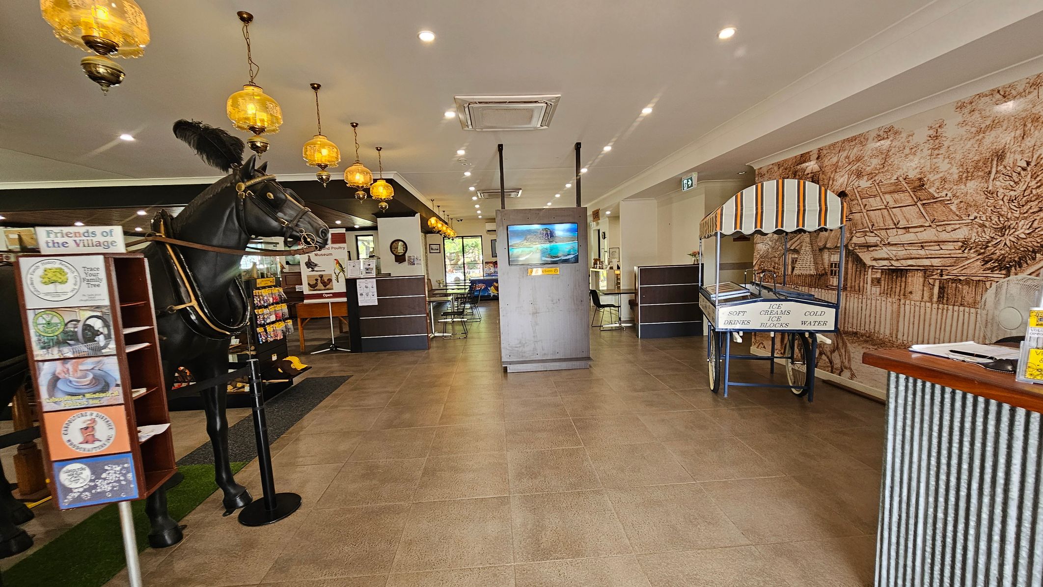 A spacious indoor area with a tiled floor and a ceiling featuring recessed lighting and hanging amber-coloured lamps. On the left, there's a large black horse statue and a display stand with brochures. In the centre, a digital screen is mounted on a grey stand. On the right, a vintage cart with a striped canopy offers soft drinks and ice creams. The background has a mural depicting a rustic village scene. Tables and chairs are visible towards the back.